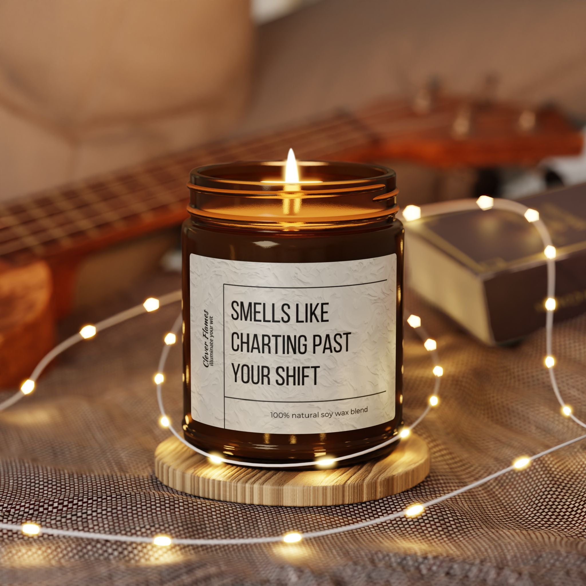 A lit candle from Printify, labeled Smells Like Charting Past Your Shift, sits on a wooden coaster surrounded by string lights. Made with a 100% natural soy wax blend and exuding relaxing lavender vibes, its ideal for Nurse Appreciation Gifts. A guitar and book are blurred in the background.