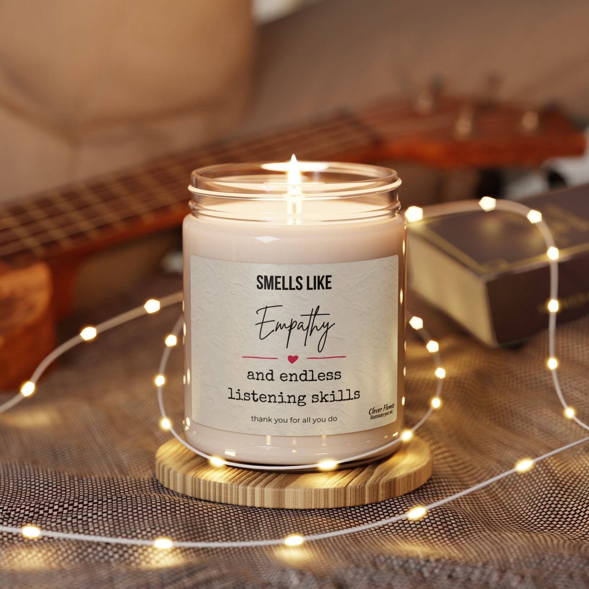 The Empathy Candle – 'Smells Like Empathy and Endless Listening Skills' 9oz Soy Candle by Printify sits in a glass jar on a wooden coaster, surrounded by string lights with a guitar and book in the background.