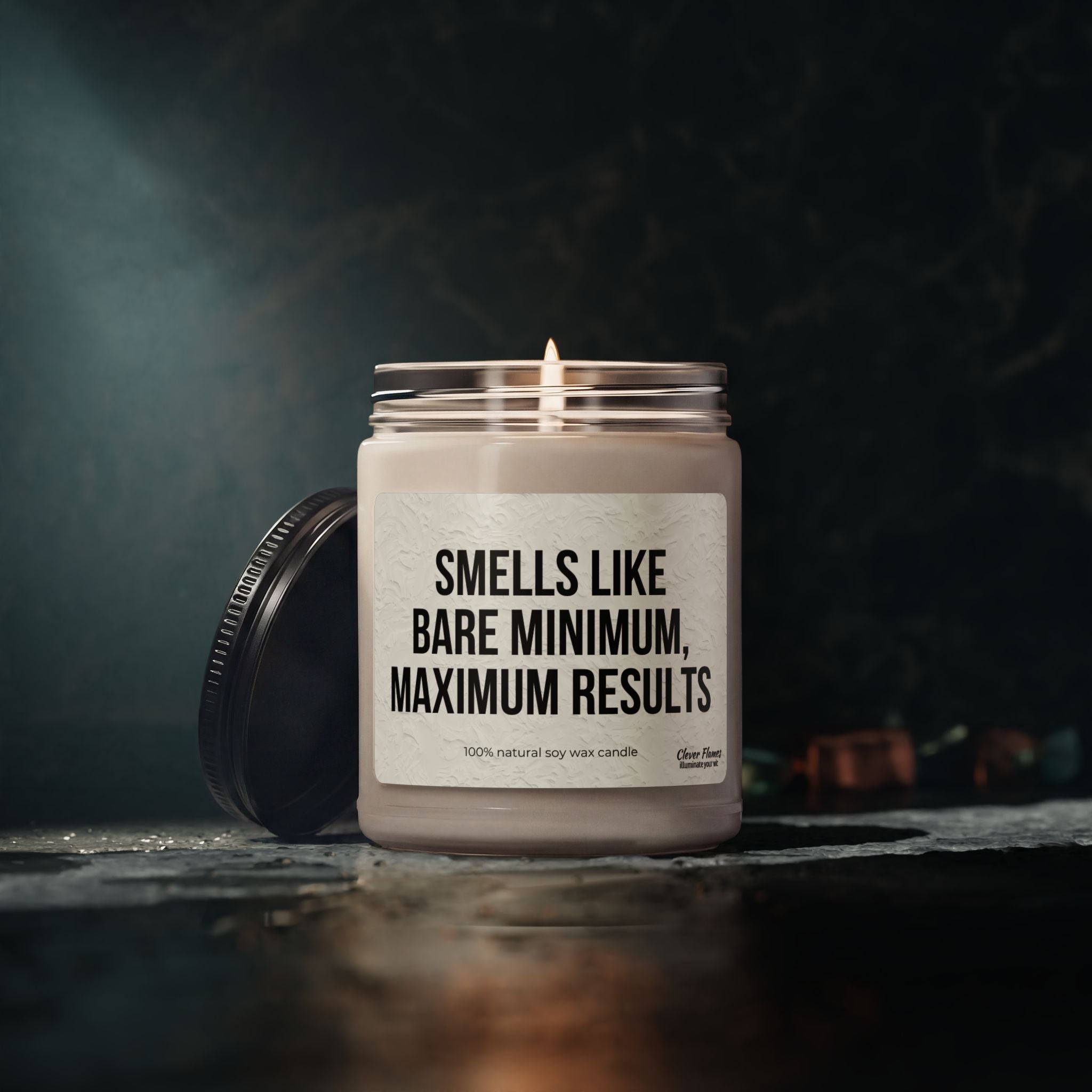 A lit 9oz Printify soy candle, "Smells Like Bare Minimum, Maximum Results," in a reusable glass jar with black lid lies on its side. The vegan, 100% natural candle sits on a dark surface with a softly blurred background.
