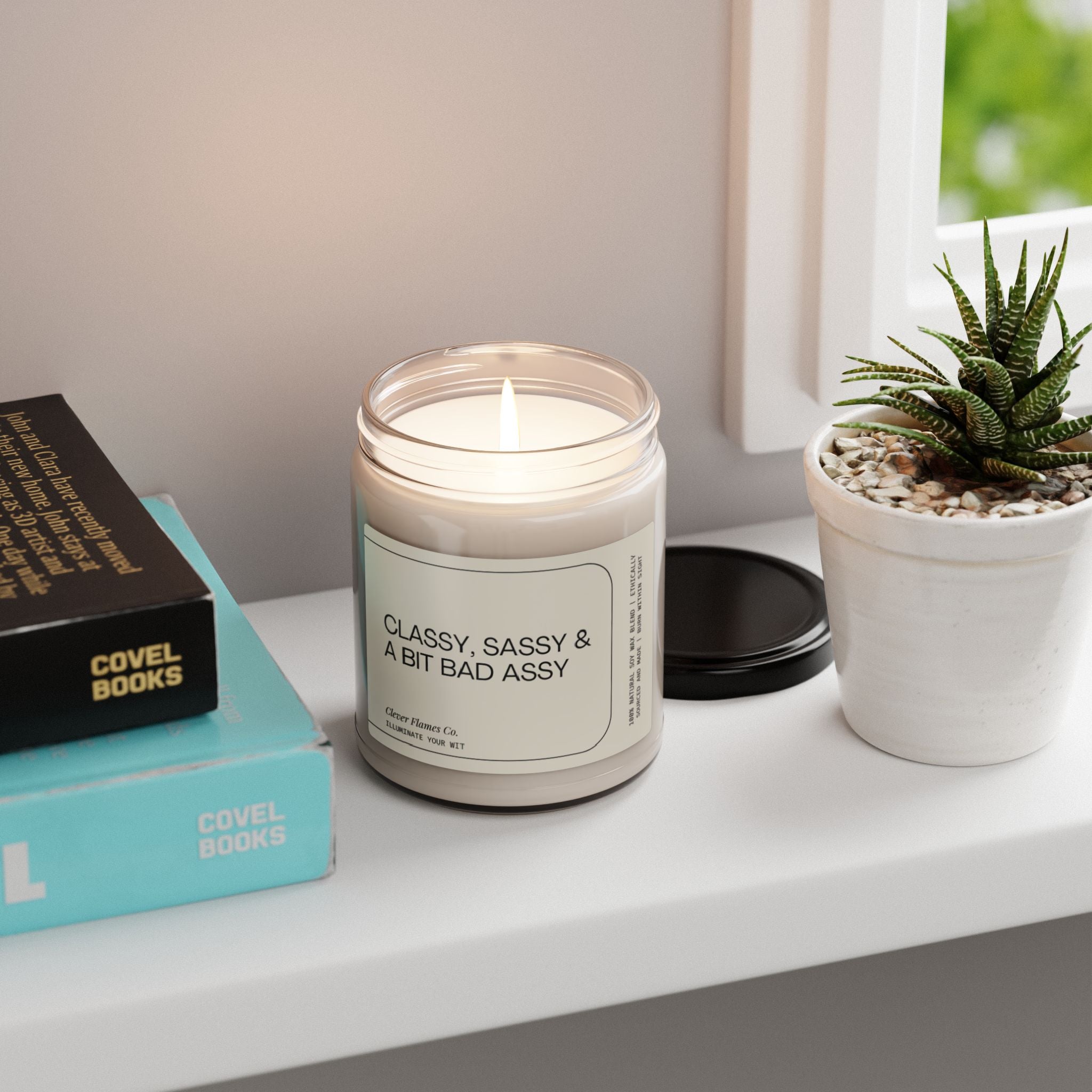 A lit Printify candle called "Classy, Sassy And Bit Bad Assy Soy Candle" made from essential oil soy wax is displayed on a white shelf next to a small potted succulent and piles of books. The aromatic scents permeate the air while a window in the background showcases lush greenery.