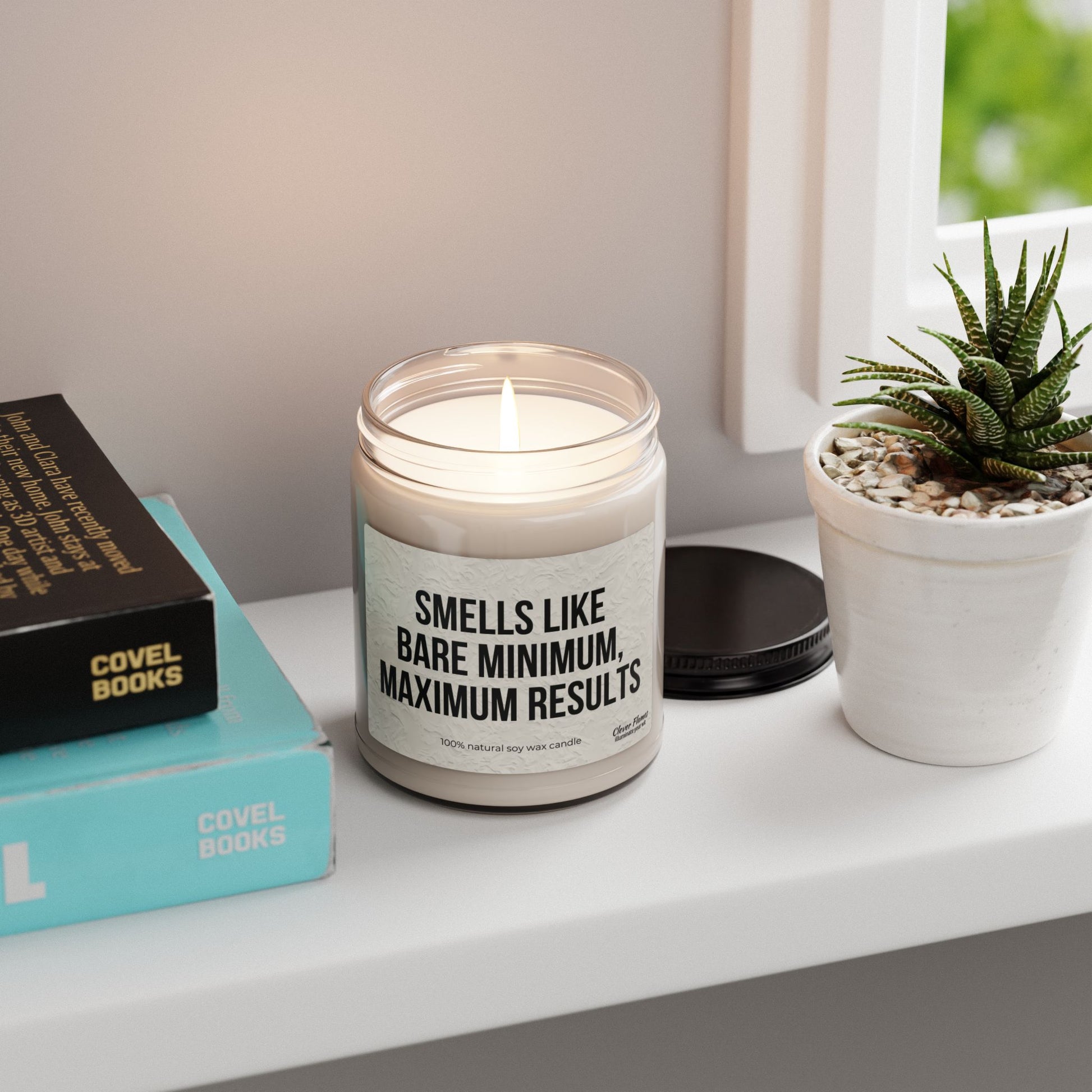 A lit Printify 9oz soy candle labeled "Smells Like Bare Minimum, Maximum Results" sits on a white shelf near a potted succulent and books, with its black lid off and resting nearby.
