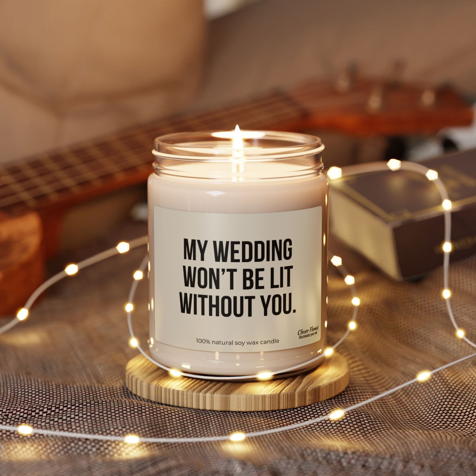 Surrounded by fairy lights, the Funny Bridesmaid Candle Proposal Idea Keepsake from Wedding features the text "My Wedding Won't Be Lit Without You." Made with eco-friendly soy wax, a ukulele and a book are softly blurred in the background, enhancing a cozy ambiance.