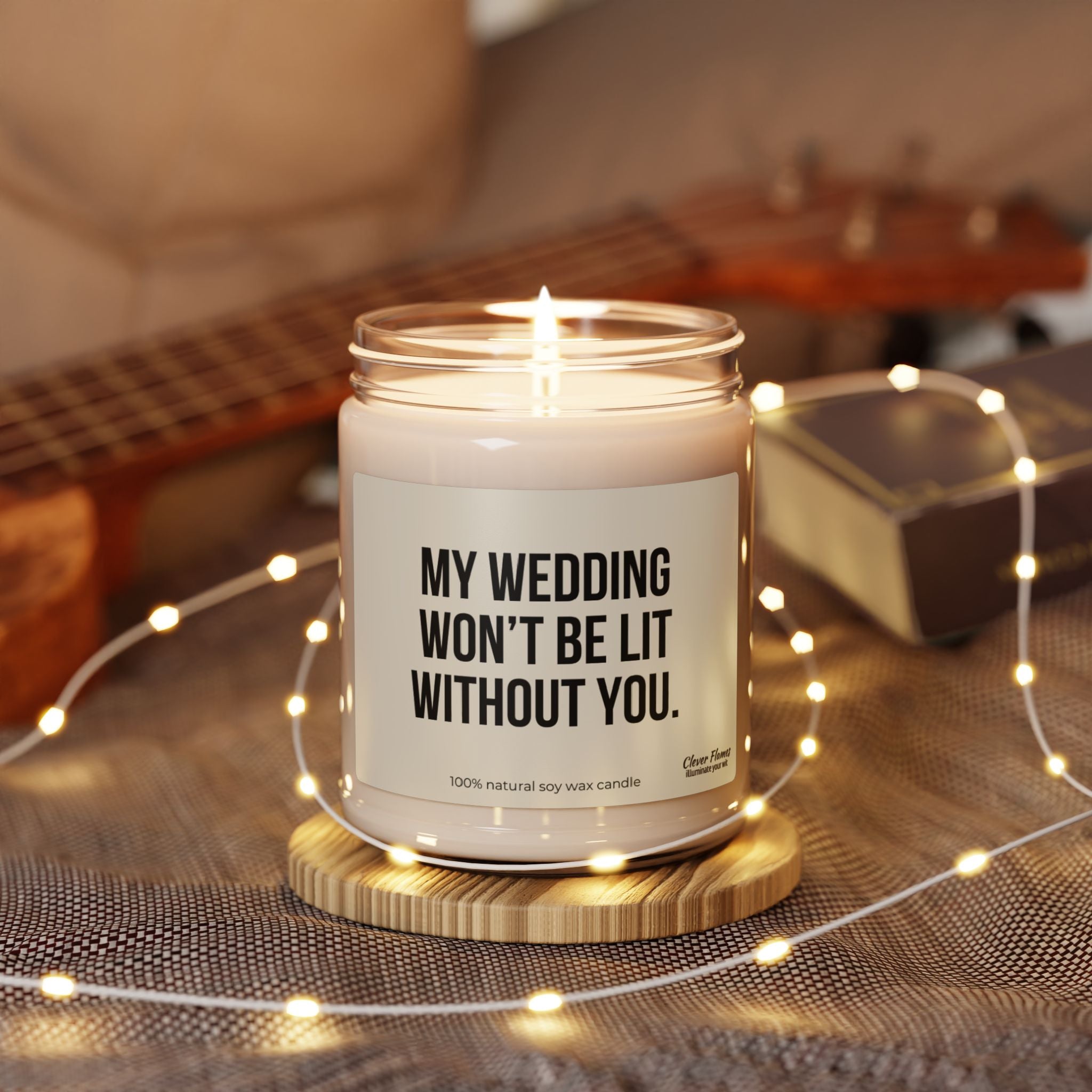 Surrounded by fairy lights, the Funny Bridesmaid Candle Proposal Idea Keepsake from Wedding features the text "My Wedding Won't Be Lit Without You." Made with eco-friendly soy wax, a ukulele and a book are softly blurred in the background, enhancing a cozy ambiance.