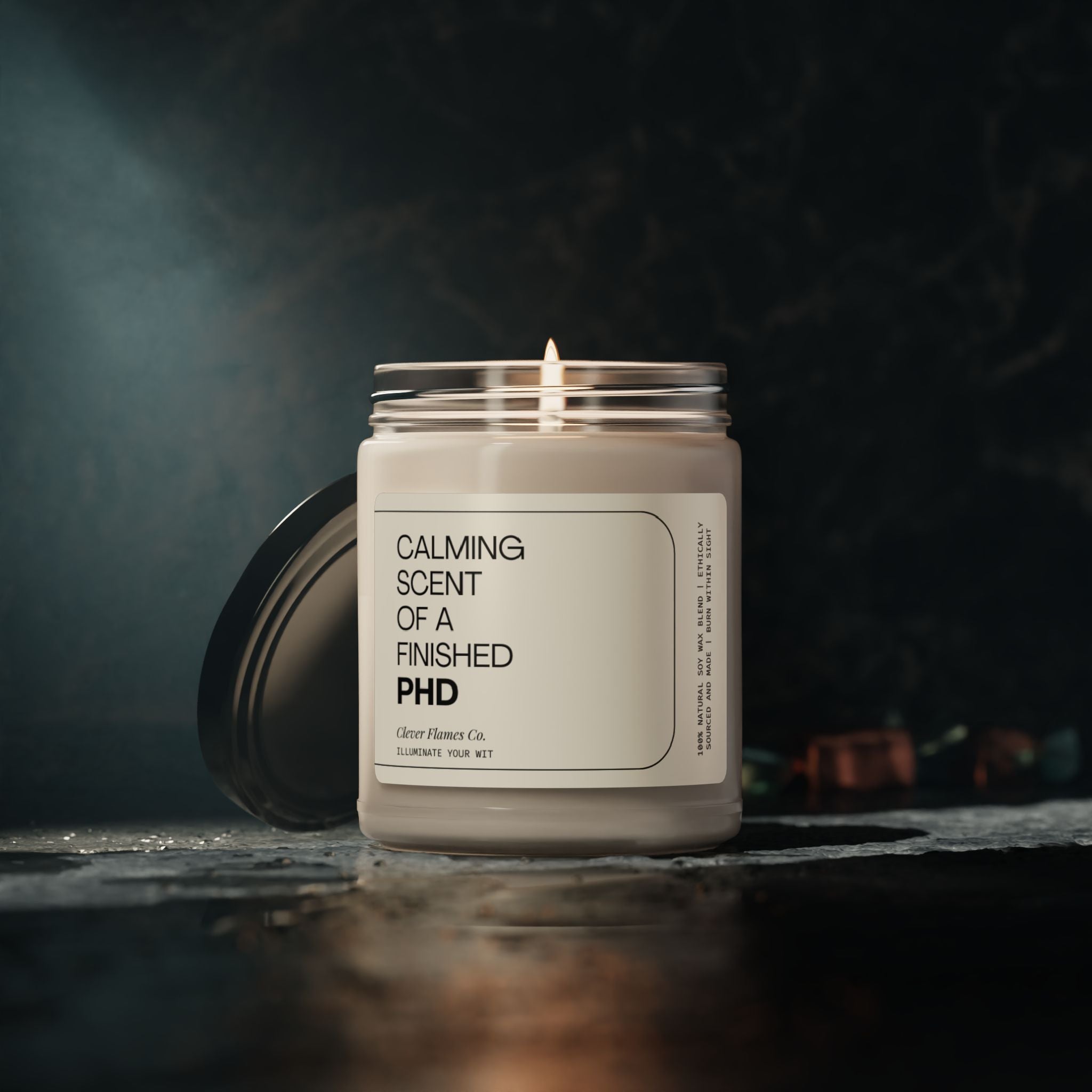 A candle labeled "Finished PhD" by Printify burns on a dark surface. The jar is open, with the lid resting beside it. This perfect graduation gift for a college grad or someone completing their master's degree casts a gentle glow in the dimly lit background, creating a tranquil atmosphere ideal for relaxation.