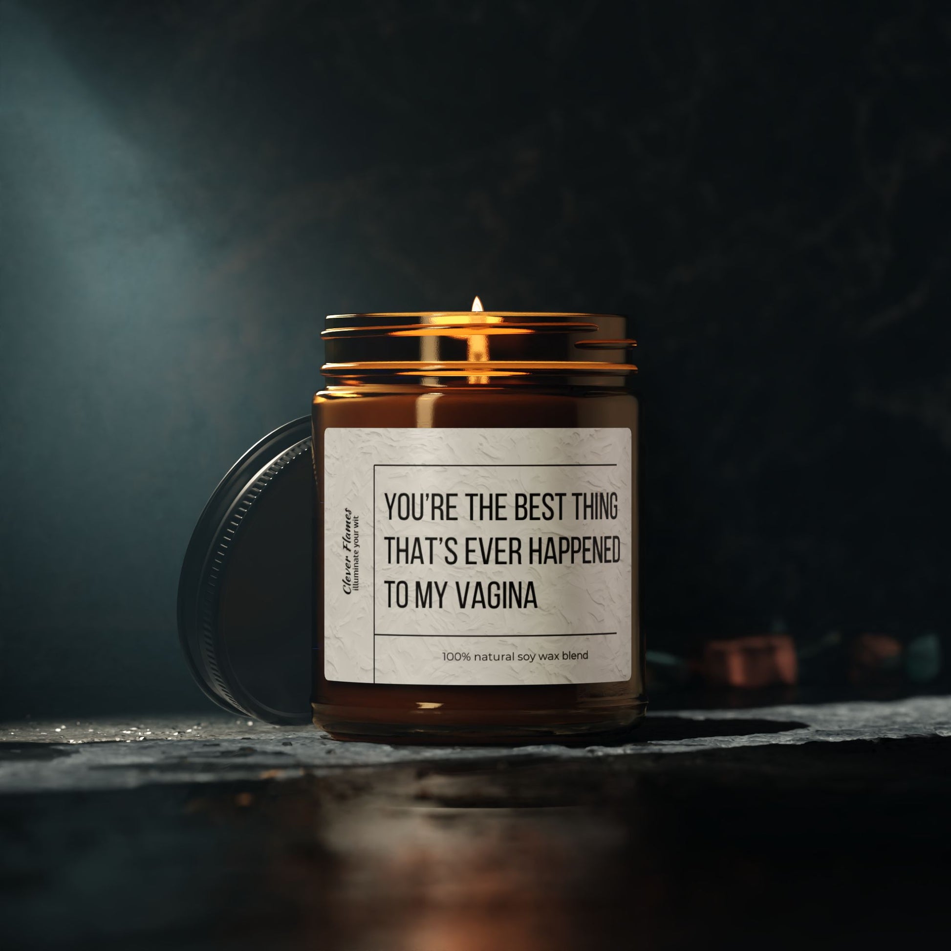 A Printify soy candle labeled Best Thing Thats Ever Happened To My Vagina, in a brown jar with a metal lid, brightens up the dark setting on a textured surface, filling the room with Christmas Warmth.