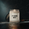 A Printify Its Giving Wifey candle, crafted from natural soy wax, rests on a dark surface with its lid beside it, emitting a warm and cozy atmosphere ideal for an engagement gift or intimate moments.