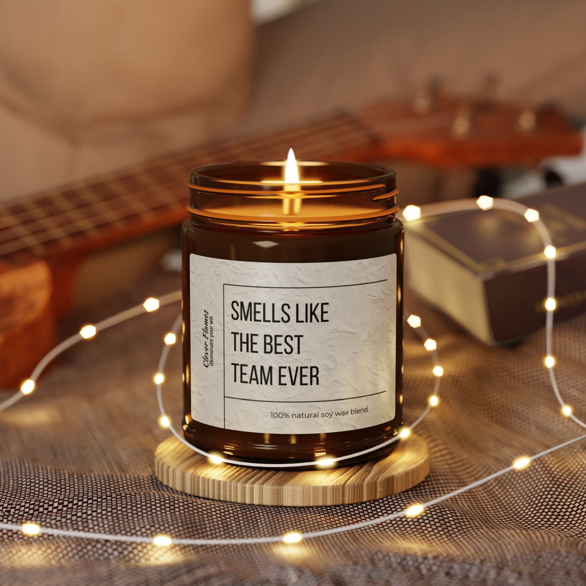 A lit brown scented soy candle from Printify, labeled "Scented Soy Candle, Best Team Employee Christmas Corporate Gift," sits on a wooden coaster. It is surrounded by glowing string lights with a blurred background featuring a ukulele and a book, making it the perfect eco-friendly corporate gift choice.