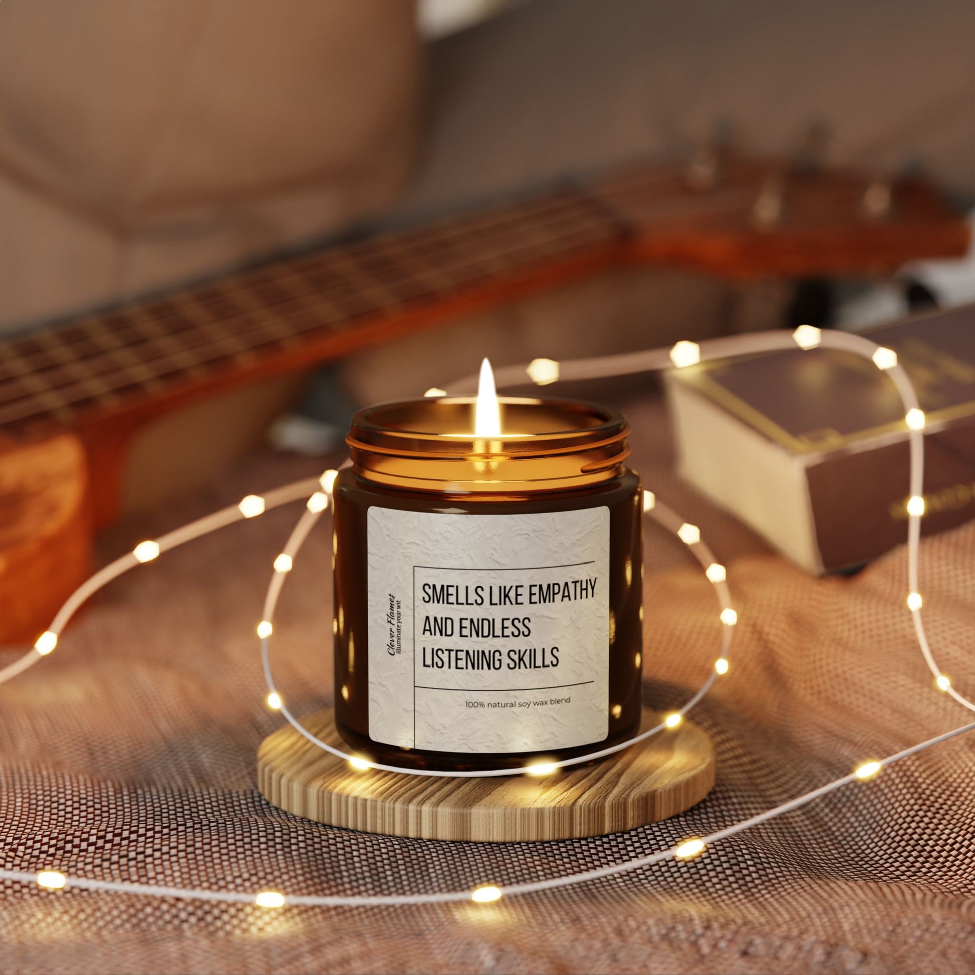 A soy candle from Printify, named the Counselor Candle with the label "Smells Like Empathy & Endless Listening Skills," sits in a brown jar on a wooden coaster, surrounded by string lights for a cozy Christmas ambiance. A guitar and closed book in the blurred background enhance its charm.