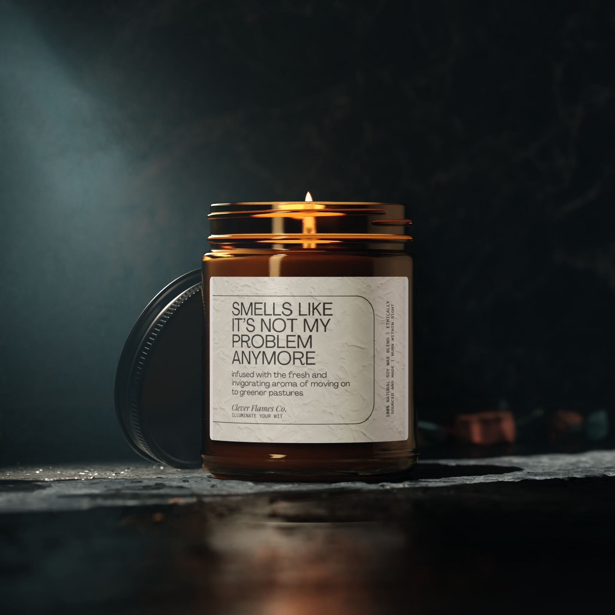 A lit brown soy candle jar from Printify's collection, labeled "Funny Retirement Scented Soy Candle - Not My Problem Anymore," rests on a dark surface, spreading the soothing Atlantis Whisper aroma. Blurred small copper objects in the background contribute to the moody ambiance, making it an ideal retirement gift.