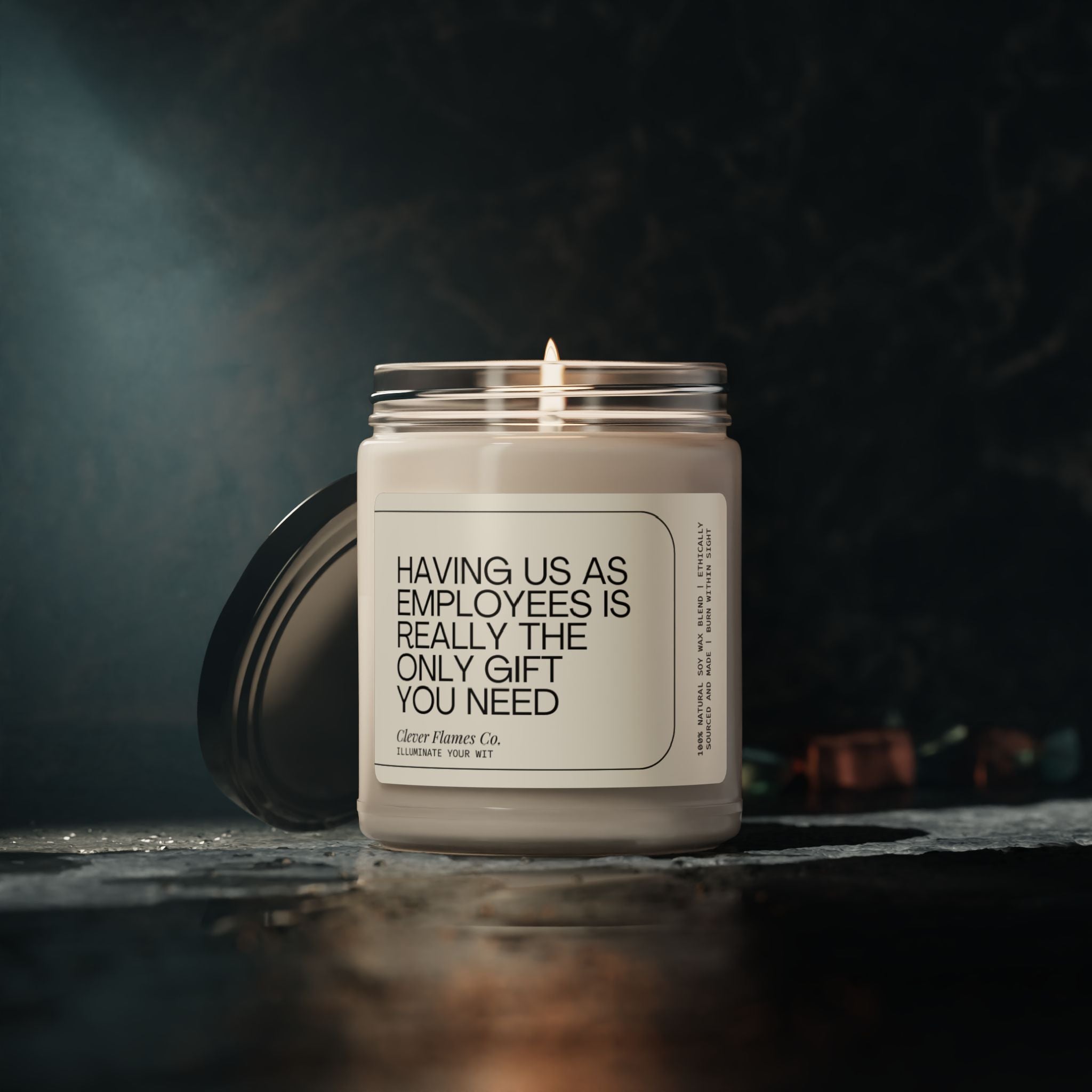 A lit Printify candle, scented with cinnamon vanilla soy wax, sits on a dark surface. The label humorously reads, "Having us as employees is really the only gift you need." With its cozy atmosphere and soft glow, this 9oz candle makes for the perfect boss appreciation gift from the collection: Funny Boss Gift Scented Soy Candle, Office Party Gift, Best Boss Ever for Boss Day.