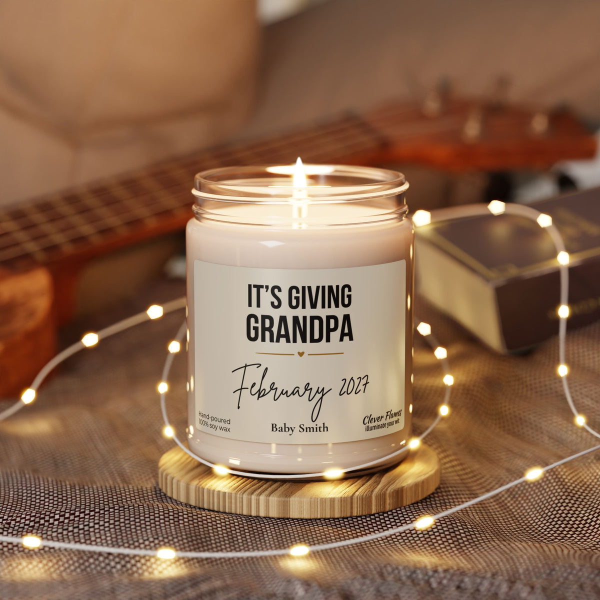 A Grandpa Pregnancy Announcement Candle in a glass jar reading Its giving Grandpa, February 2027, Baby Smith, sitting on a wooden coaster surrounded by string lights.