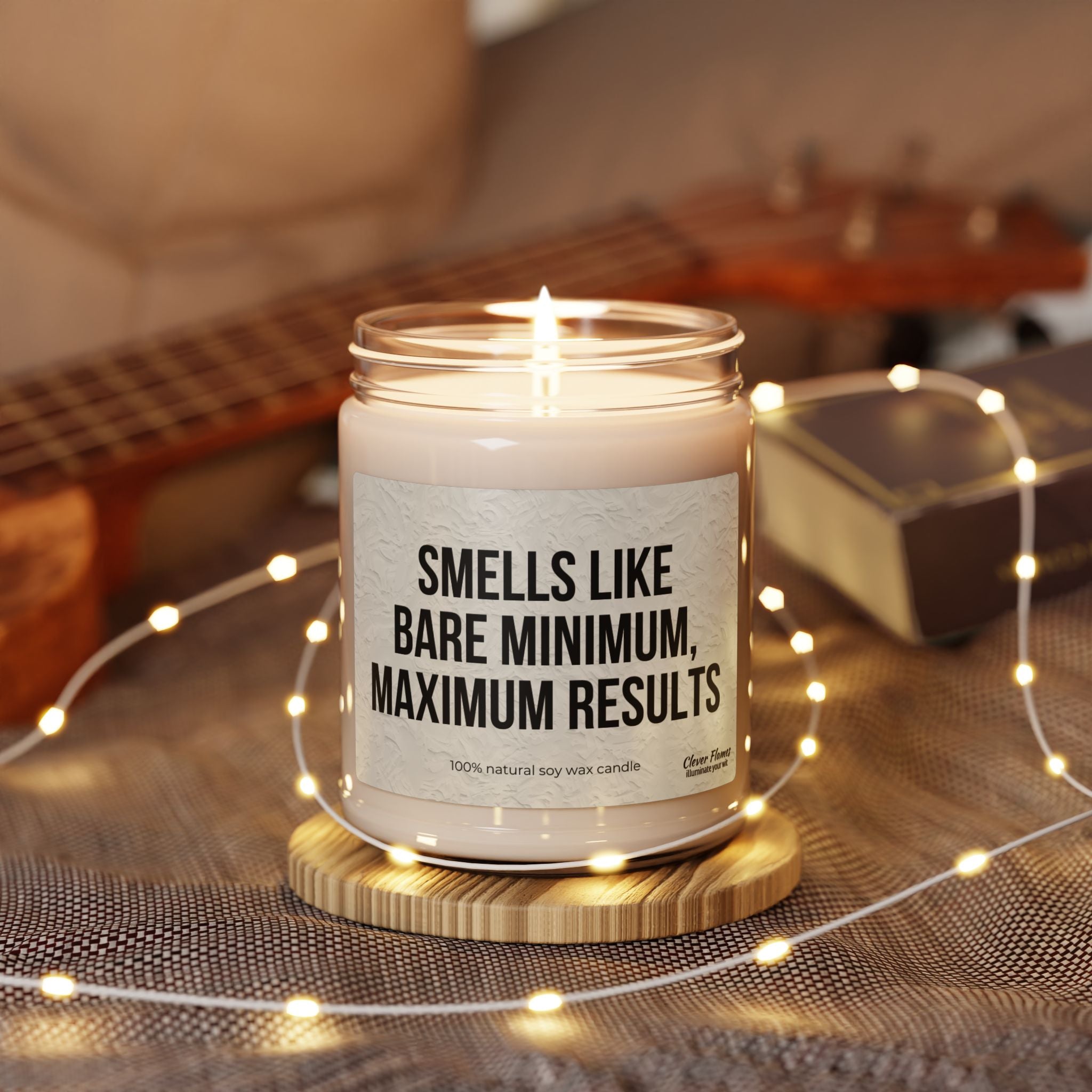 Printify's 9oz Soy Candle—"Smells Like Bare Minimum, Maximum Results"—comes in a reusable glass jar on a wooden coaster, surrounded by string lights, with a guitar and closed book in the background.