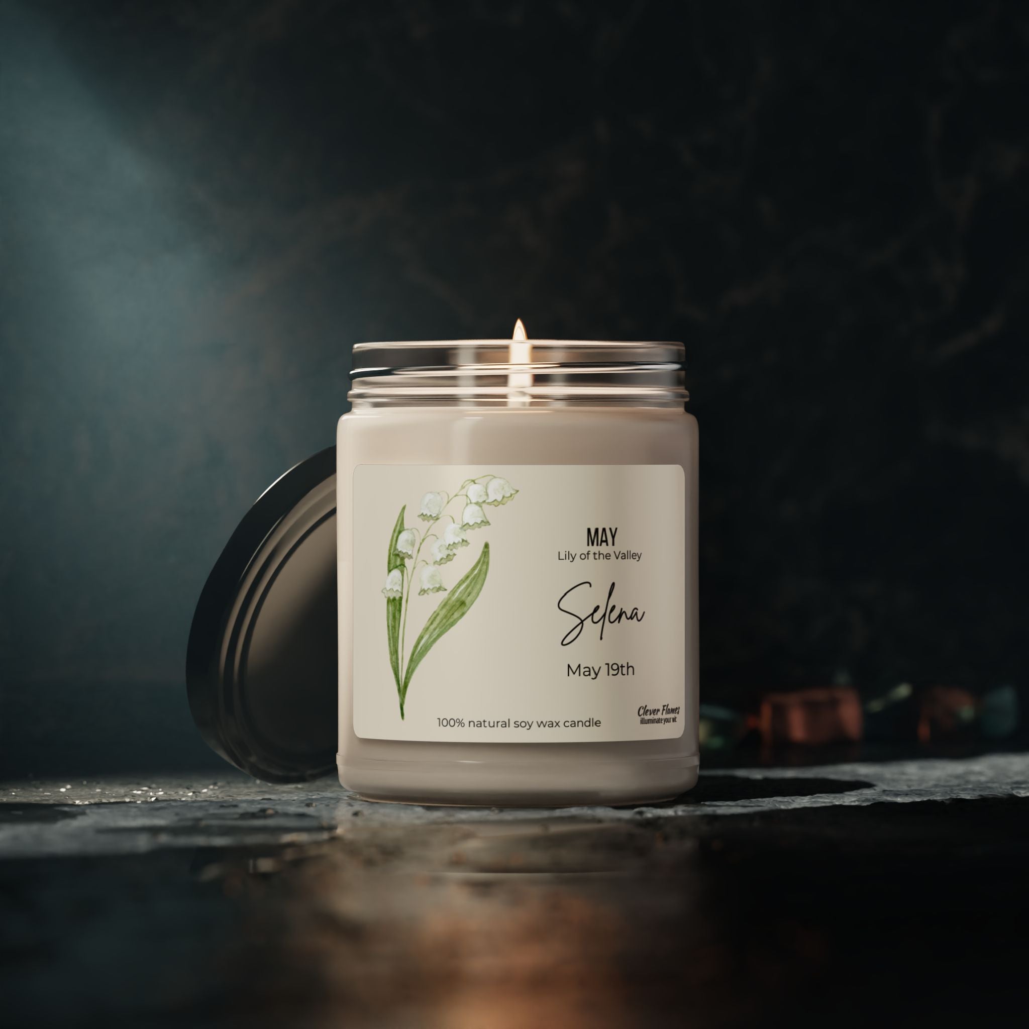 The Printify Personalized Birth Flower Candle is made of natural soy wax, featuring a May lily of the valley label with Selena and May 19th. Against a dark, blurred backdrop, it casts a cozy glow—an elegant tribute to springs essence.