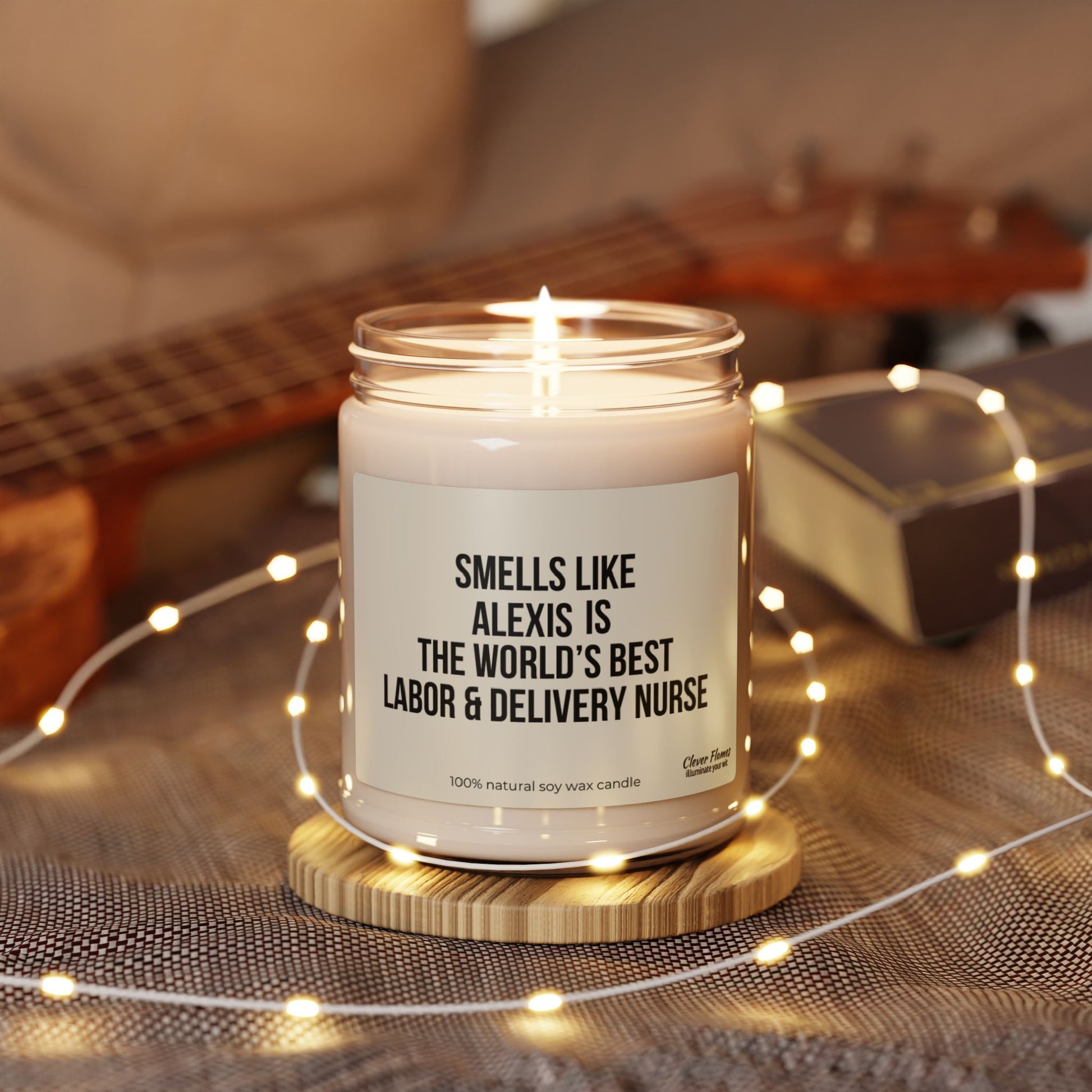 A Custom Labor and Delivery Nurse Candle from Printify, personalized with the label "Smells Like Alexis is the World's Best Labor & Delivery Nurse," is beautifully surrounded by string lights. A guitar rests in the background on a fabric surface, making it an ideal nursing school gift or thank you gesture for a nurse.