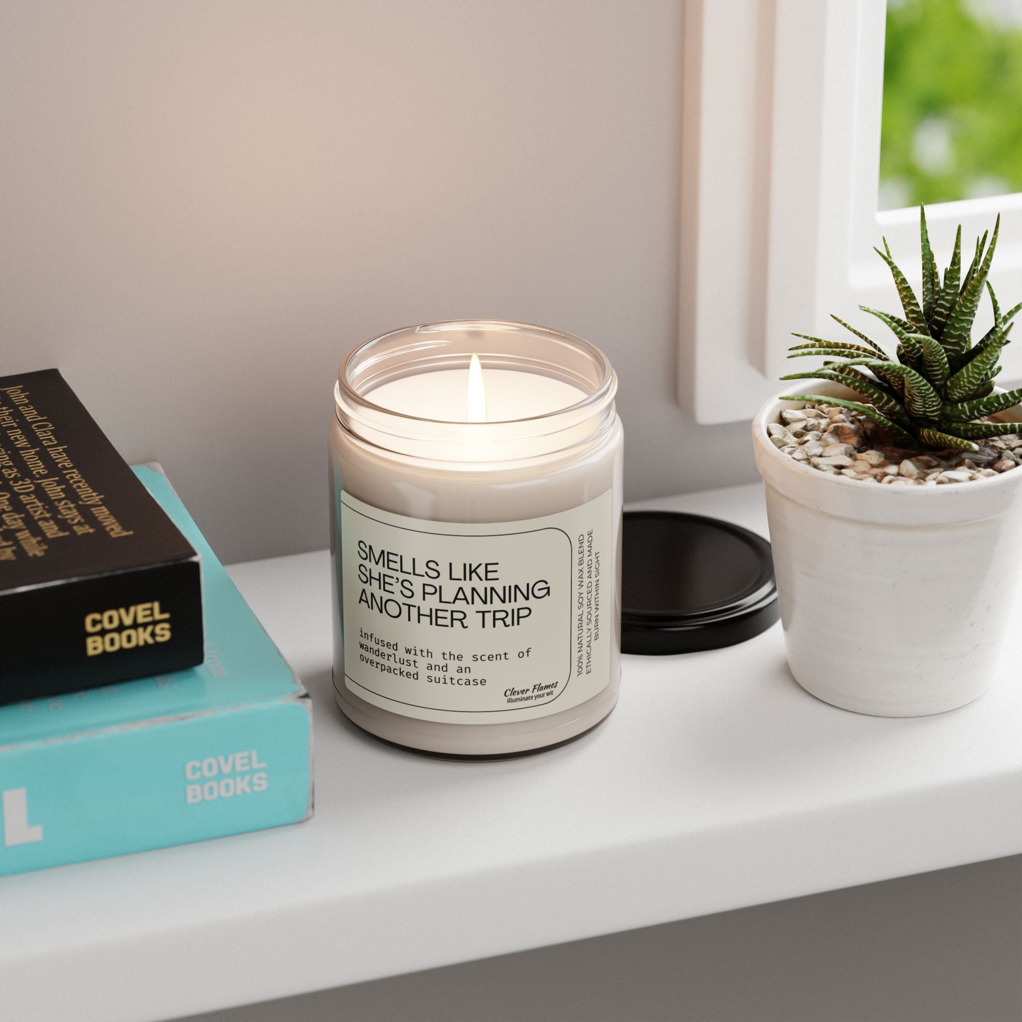A Printify 9oz "Smells Like She's Planning a Trip" soy candle sits on a white surface next to its black lid, two stacked books, and a small potted succulent.