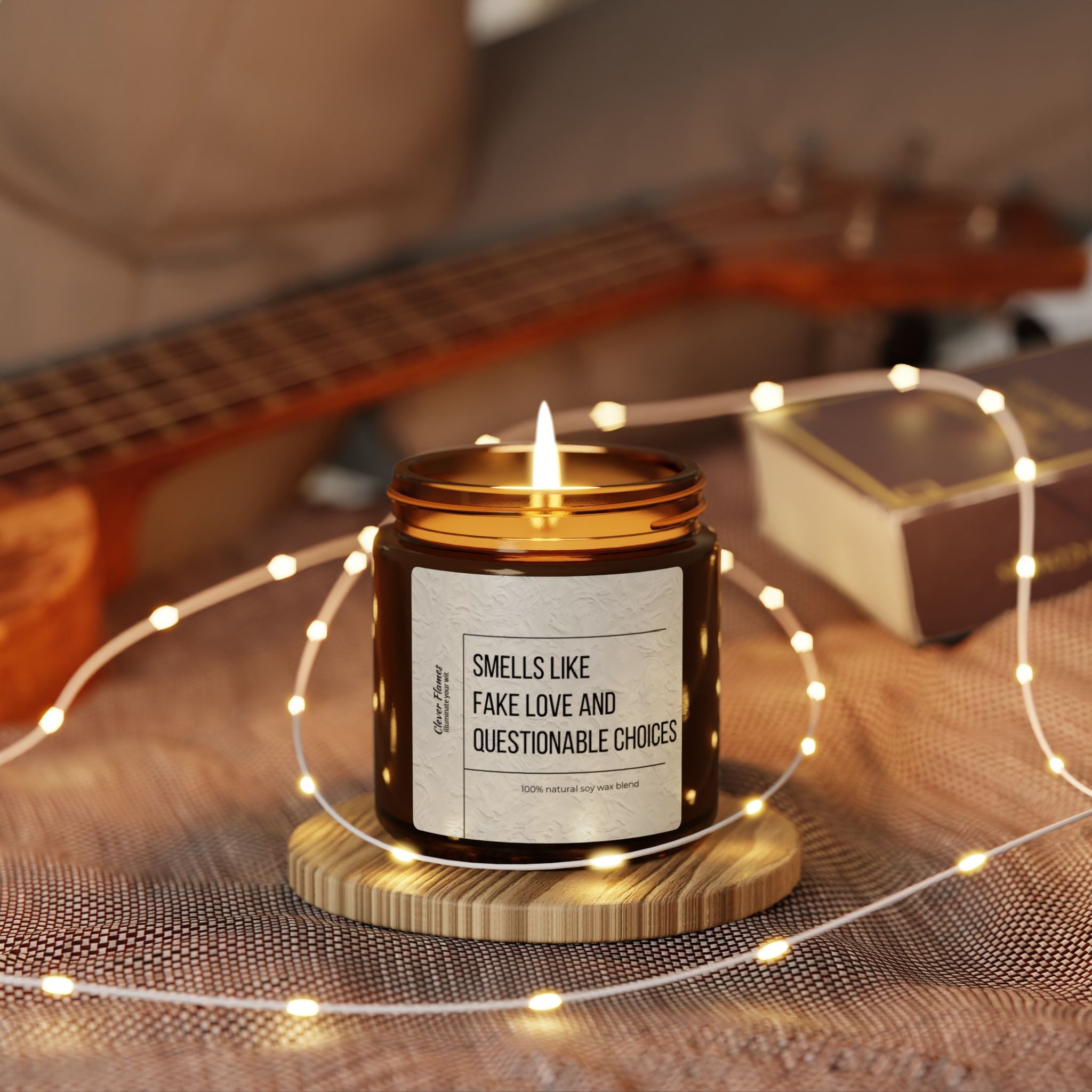 A cozy ambiance is created by the glow of a Printify Smells Like Fake Love and Questionable Choices scented soy candle, housed in an amber jar, resting on a wooden coaster. Surrounded by soft fairy lights and accompanied by a guitar and box in the background, this setting makes for an ideal romantic gift.