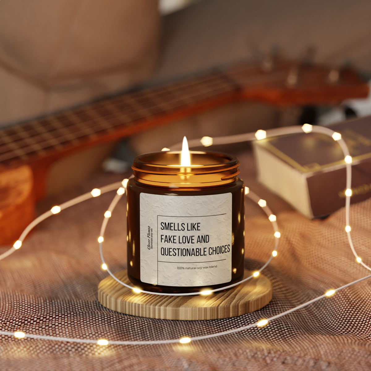A cozy ambiance is created by the glow of a Printify Smells Like Fake Love and Questionable Choices scented soy candle, housed in an amber jar, resting on a wooden coaster. Surrounded by soft fairy lights and accompanied by a guitar and box in the background, this setting makes for an ideal romantic gift.