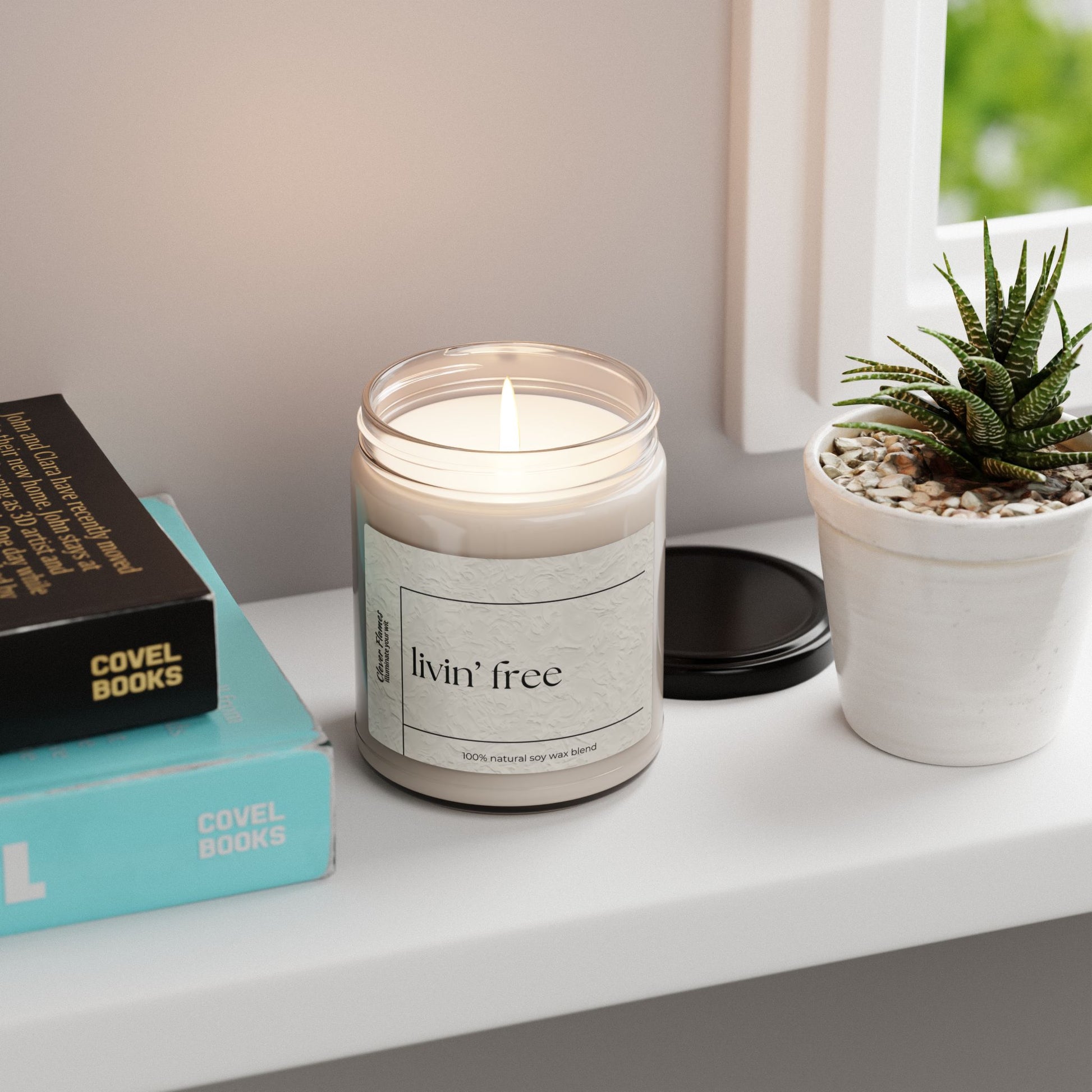 A Livin’ Free Summer Break Candle by Printify sits in a glass jar on a white windowsill beside a potted succulent and two stacked books with teal and yellow covers, perfect as a funny teacher appreciation gift.