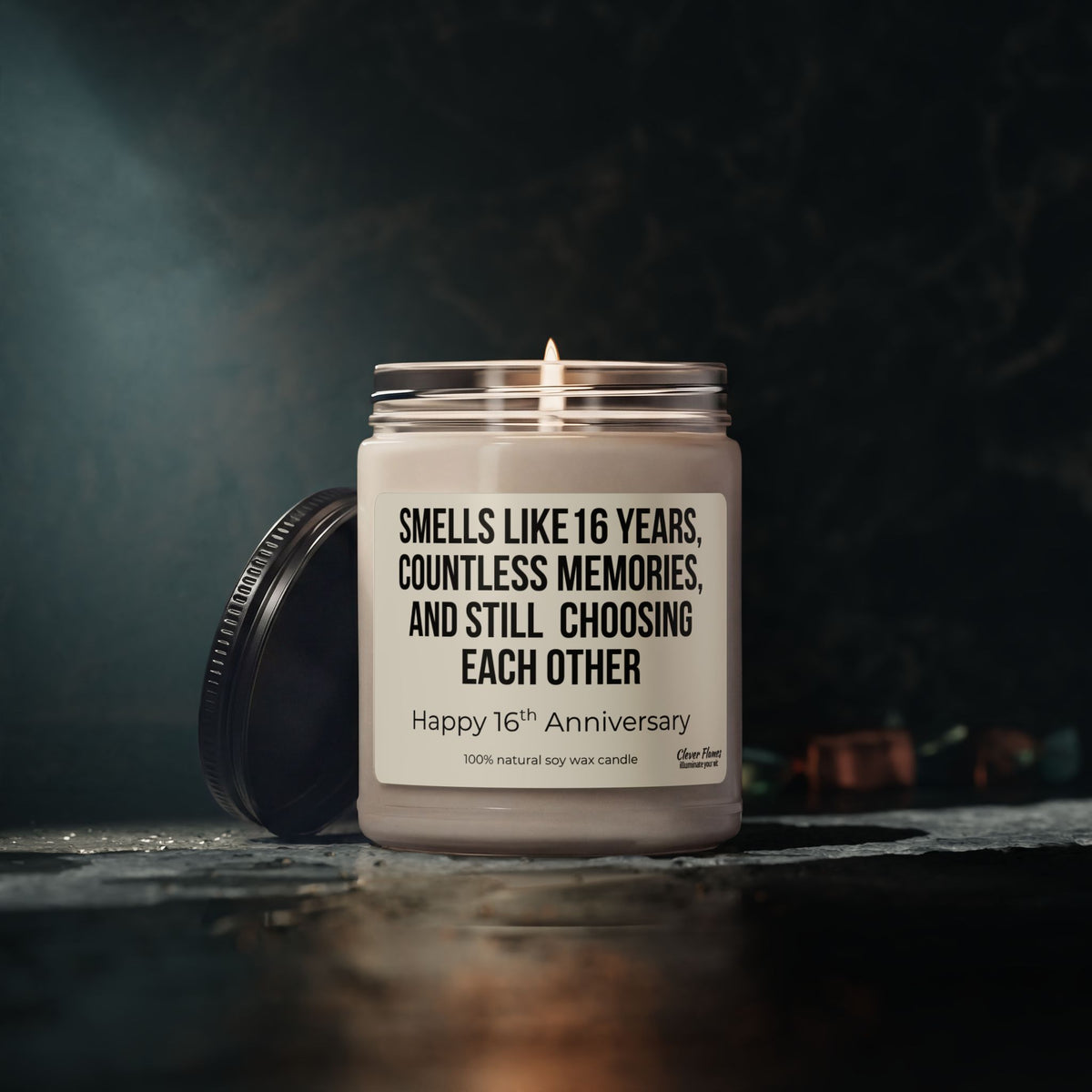 A lit white candle from Printify sits in a jar on a dark surface. The label reads: Smells like 16 years, countless memories, and still choosing each other. Happy 16th Anniversary. This 16th Anniversary Candle is made from 100% natural soy wax and makes the perfect wax anniversary gift for your wife or husband, or a thoughtful way to celebrate being married for 16 years.
