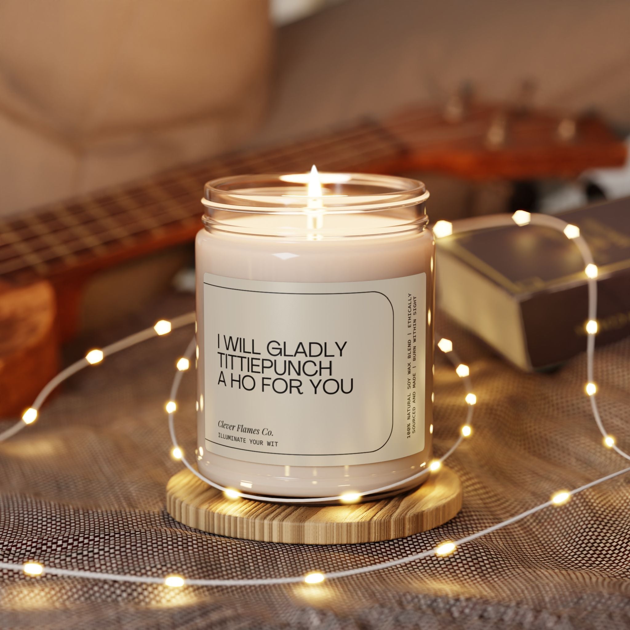 The "I Will Gladly Tittiepunch a Ho For You" soy candle by Printify, featuring an oil scent and presented in a jar with a comedic label, is lit on a wooden coaster amidst the warm glow of fairy lights. With a guitar and book subtly blurred in the backdrop, this scene creates an inviting atmosphere—making it an ideal quirky gift for her or as funny gesture for your BFF.