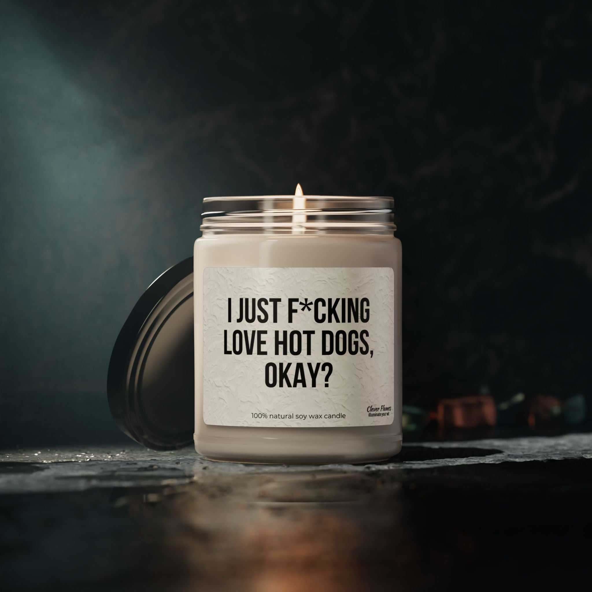 The I Just F*cking Love Hot Dogs Okay? Candle by Printify is a 9oz scented soy candle in a reusable glass jar, featuring its bold label. The candle is shown lit on a dark surface with the lid nearby and a blurred dark background.