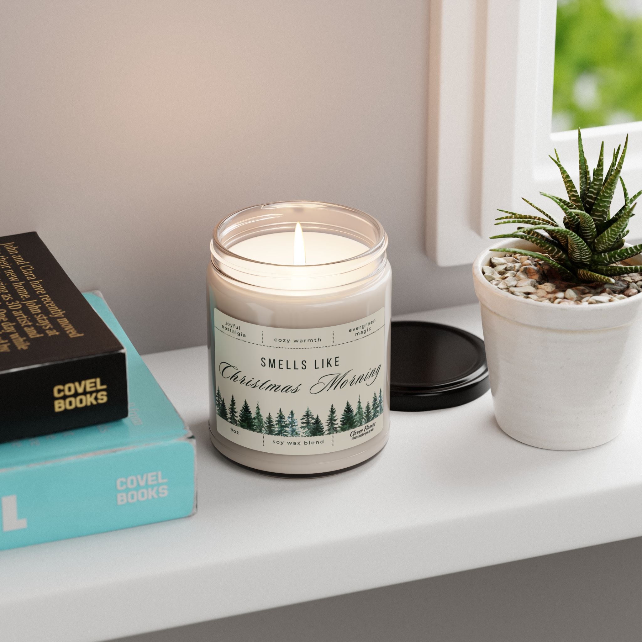 The Smells Like Christmas Morning – Holiday Soy Candle (9oz) by Printify sits on a white surface by a potted succulent, stacked books, and a window. Its black lid rests nearby, filling the cozy space with festive fragrance.