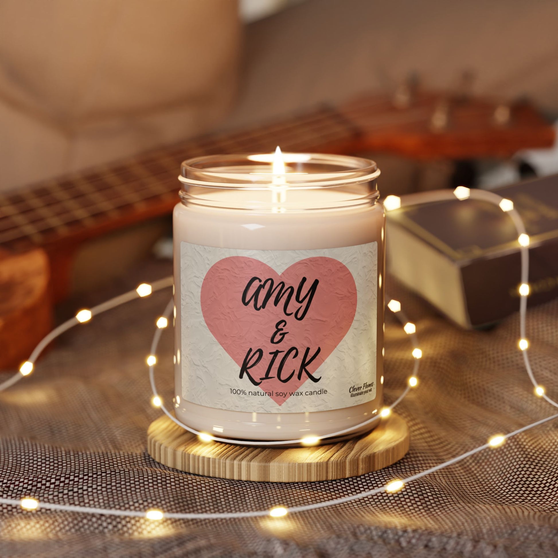 The Printify Personalized Couples Candle features a pink heart label with Amy & Rick on a jar of natural soy wax, set on a wooden coaster with string lights. This romantic keepsake makes an ideal gift, enhancing coziness alongside blurred guitars and books in the background.