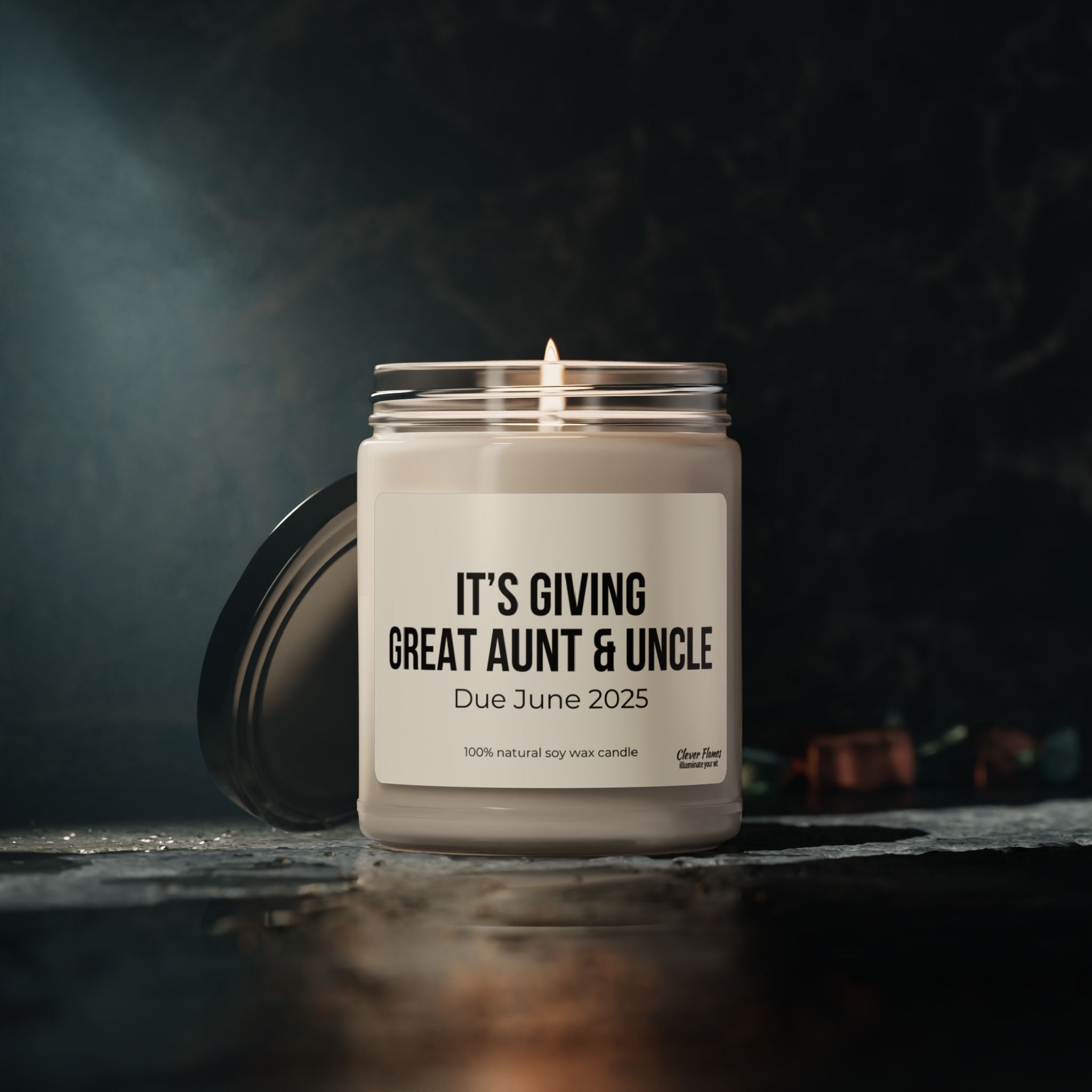 A personalized candle from Printify, featuring soy wax in a glass jar, emits a warm glow. The label reads "Great Aunt and Uncle Pregnancy Announcement Candle Due June 2025," beautifully setting an intimate scene with its dark background, leaf accents, and wooden surface. This baby announcement gift is the perfect way to reveal your joyful news to a great aunt and uncle.