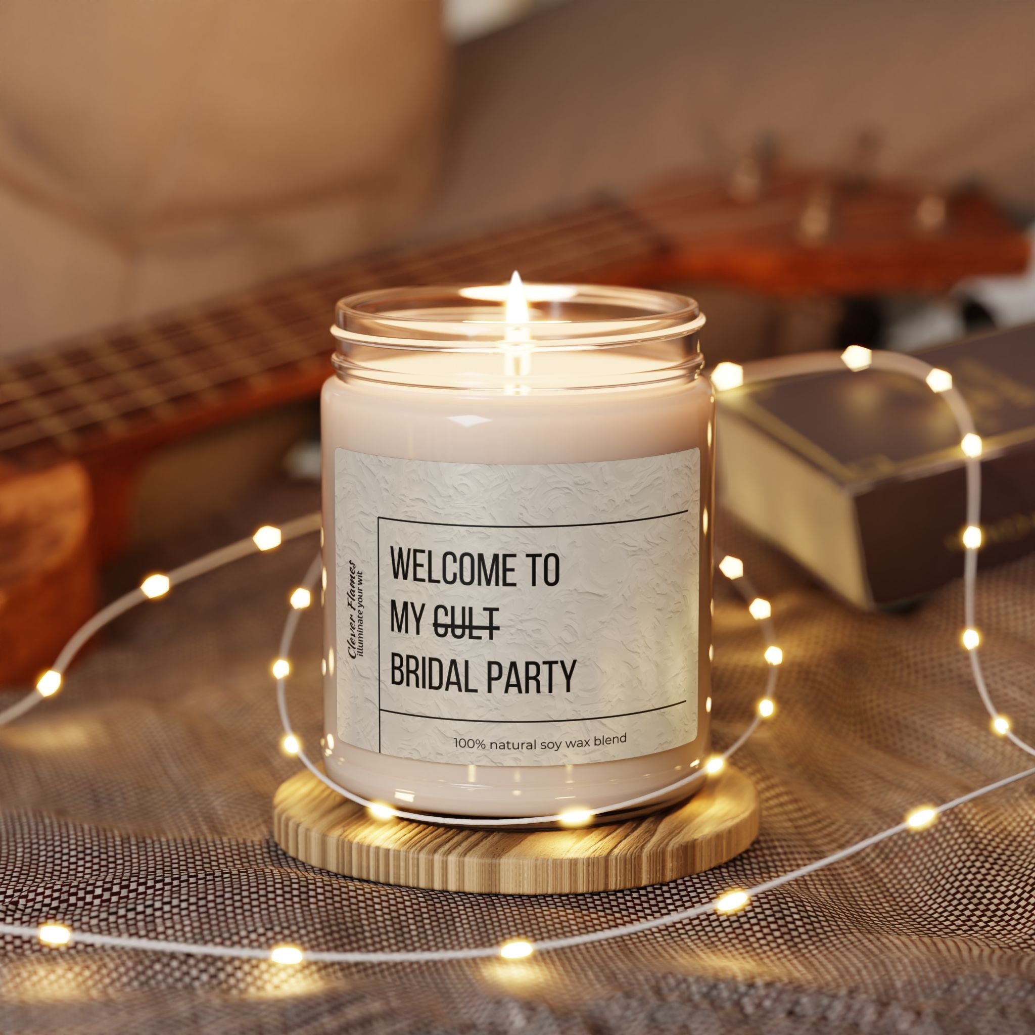 The Printify Welcome to My Cult Bridal Party Scented Soy Candle (9oz) glows in a glass jar on a wooden coaster, with warm string lights and a blurred ukulele in the background creating a cozy, aromatic vibe.