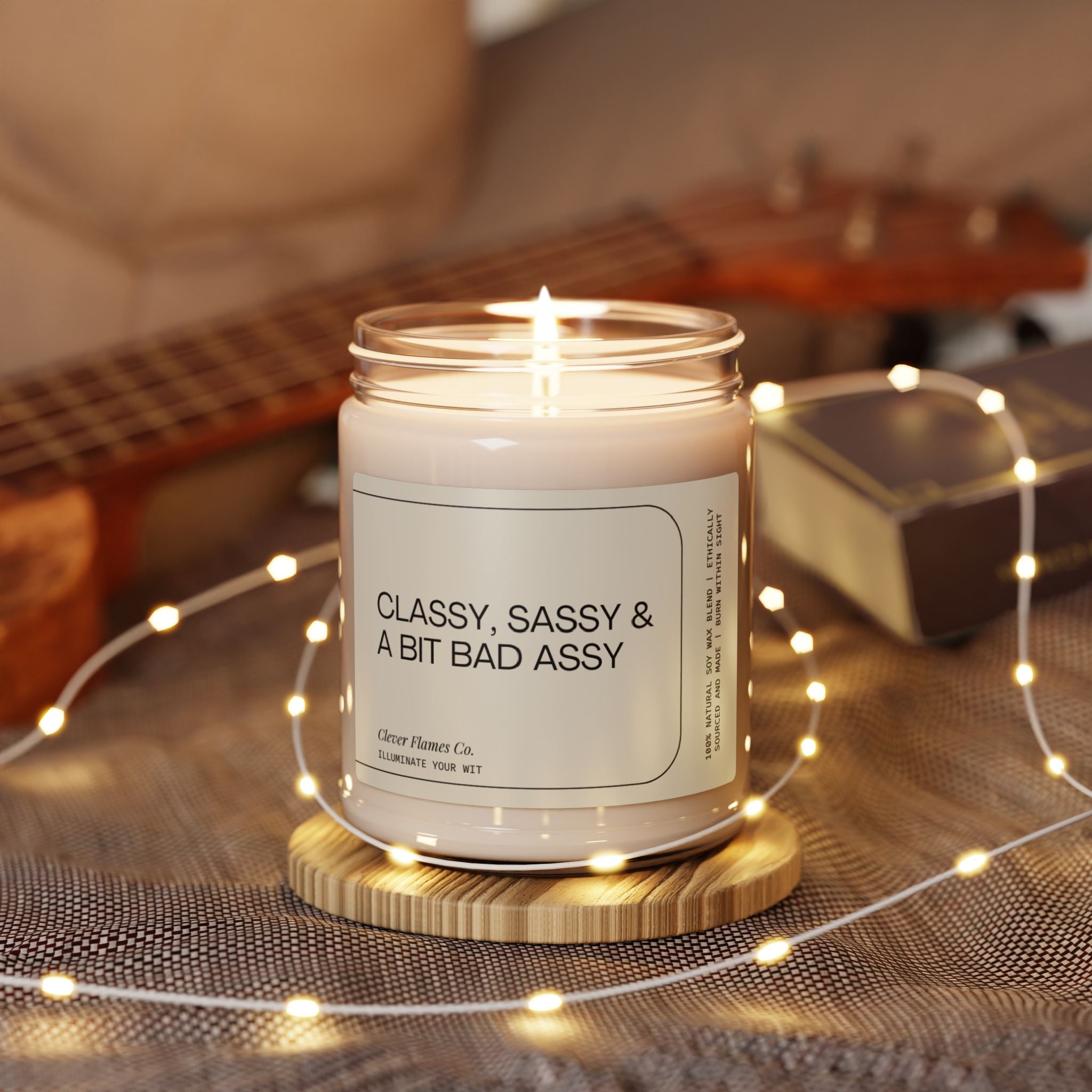 A lit Printify soy candle labeled "Classy, Sassy And Bit Bad Assy" crafted from natural soy wax sits on a wooden coaster surrounded by tiny lights. In the background, a ukulele and a book softly blur into focus.