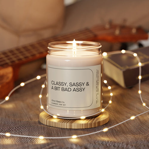 A lit Printify soy candle labeled "Classy, Sassy And Bit Bad Assy" crafted from natural soy wax sits on a wooden coaster surrounded by tiny lights. In the background, a ukulele and a book softly blur into focus.