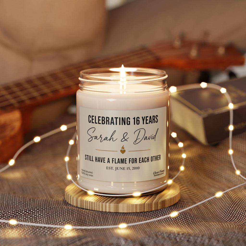 A Personalized 16th Anniversary Candle from Printify, custom-labeled with “Celebrating 16 years, Sarah & David. Still have a flame for each other. Est. June 15, 2018,” rests on a wooden base encircled by string lights with a guitar in the background—an elegant and meaningful Custom Wax Anniversary Gift for Husband or Wife, perfect for commemorating 16 years of lasting love.