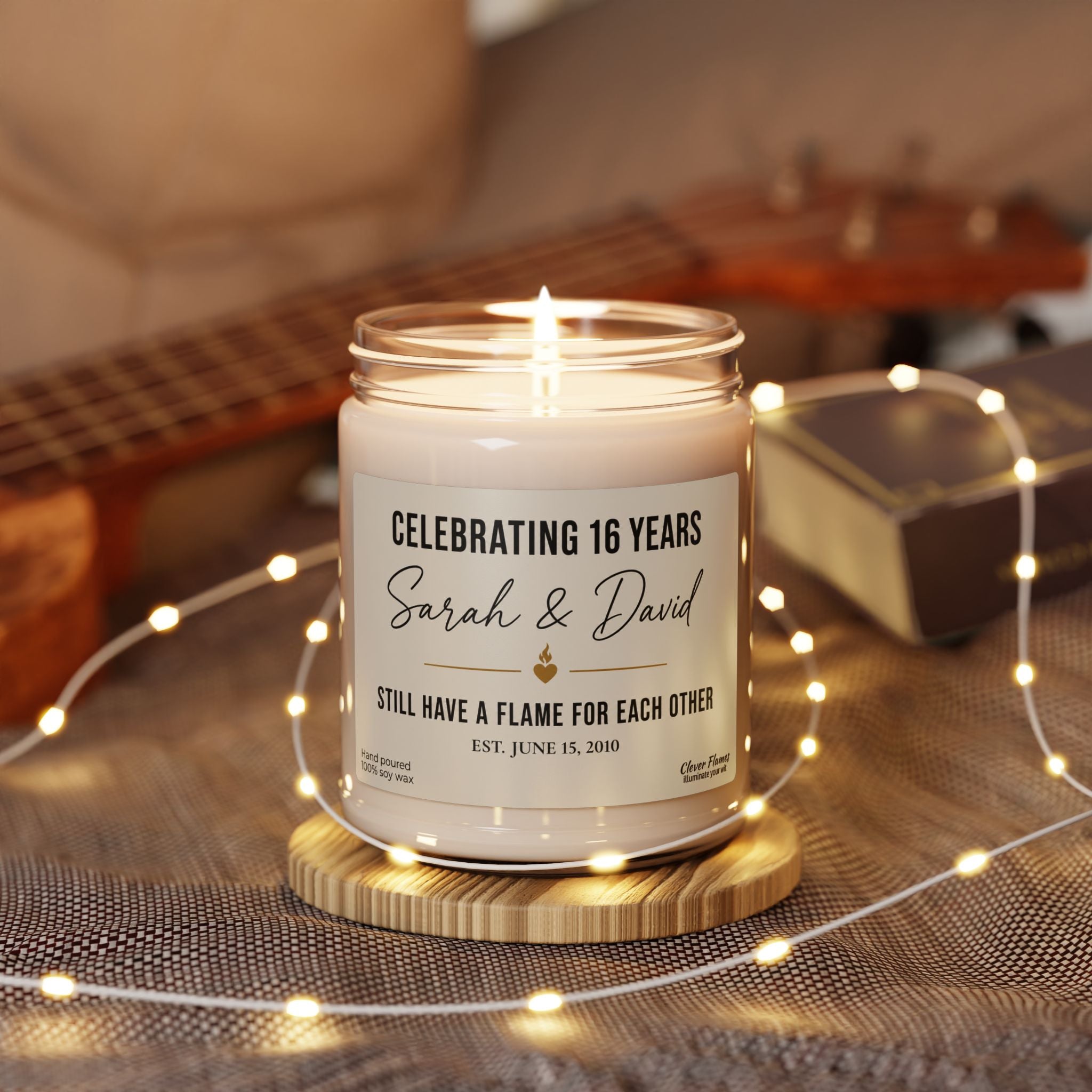 A Personalized 16th Anniversary Candle from Printify, custom-labeled with “Celebrating 16 years, Sarah & David. Still have a flame for each other. Est. June 15, 2018,” rests on a wooden base encircled by string lights with a guitar in the background—an elegant and meaningful Custom Wax Anniversary Gift for Husband or Wife, perfect for commemorating 16 years of lasting love.