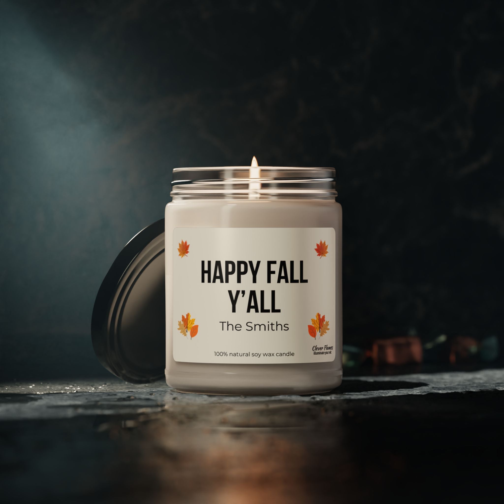 A personalized scented soy candle from Printify, featuring the label "Happy Fall Yall" and "The Smiths," encased in a glass jar adorned with autumn leaf designs. This jar emanates cozy fall vibes against a dark, moody background—a perfect decor piece for snug autumn evenings.