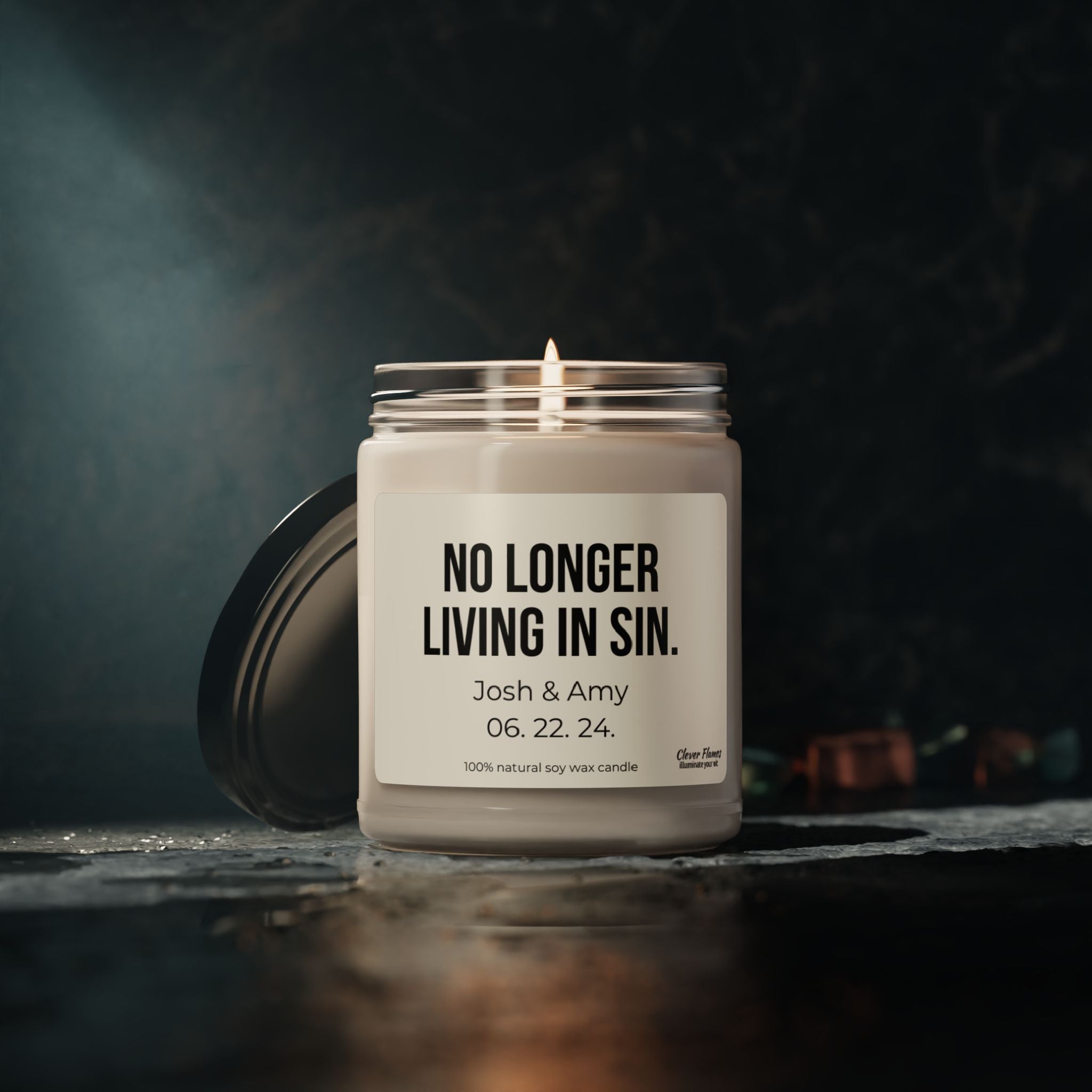 A soy candle from the Wedding brand sits on a dark surface. The label reads: NO LONGER LIVING IN SIN, Josh & Amy 06. 22. 24. Crafted from 100% natural soy wax, this personalized gift offers a gentle glow—making it an ideal anniversary present to celebrate special moments together.