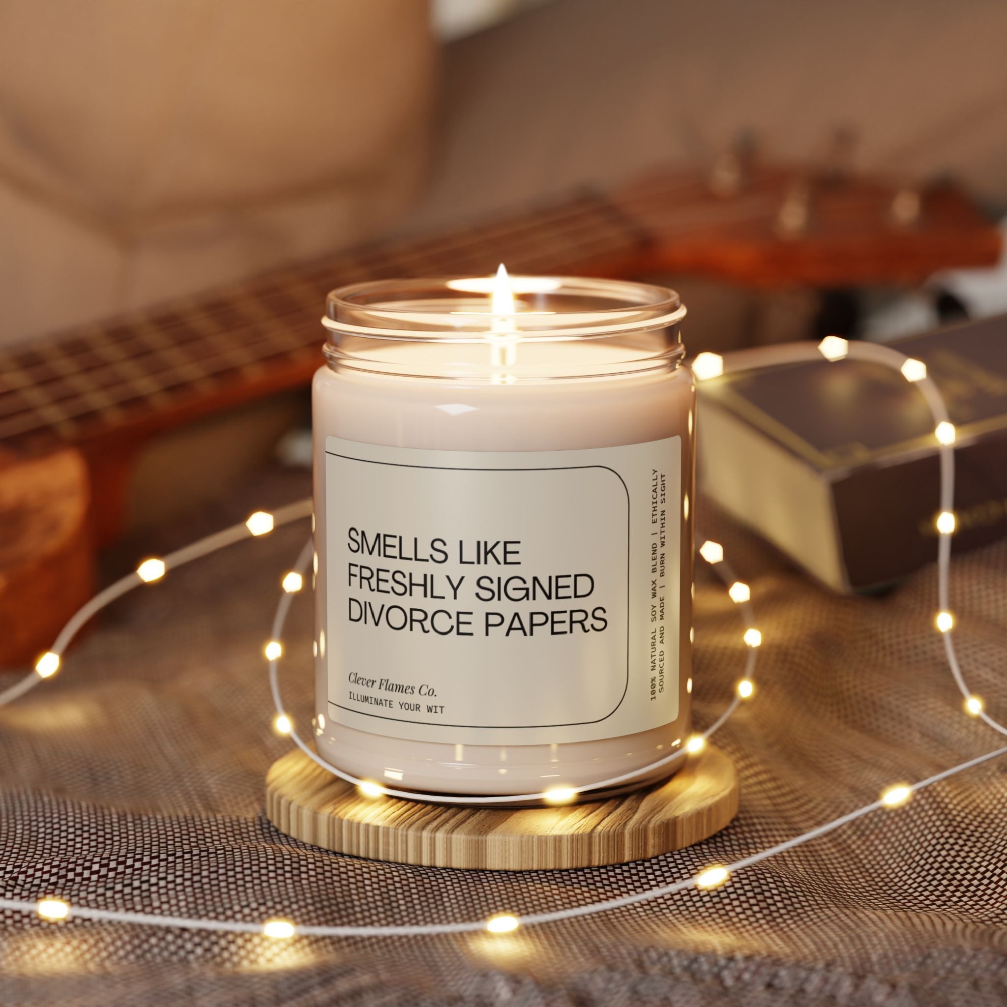 A candle from Printify, named "Smells Like Freshly Signed Divorce Papers," rests elegantly in a glass jar crafted from natural soy wax, set upon a wooden coaster. Encircled by twinkling string lights with a guitar and book in the background, this unique piece makes for the perfect funny divorce gift.