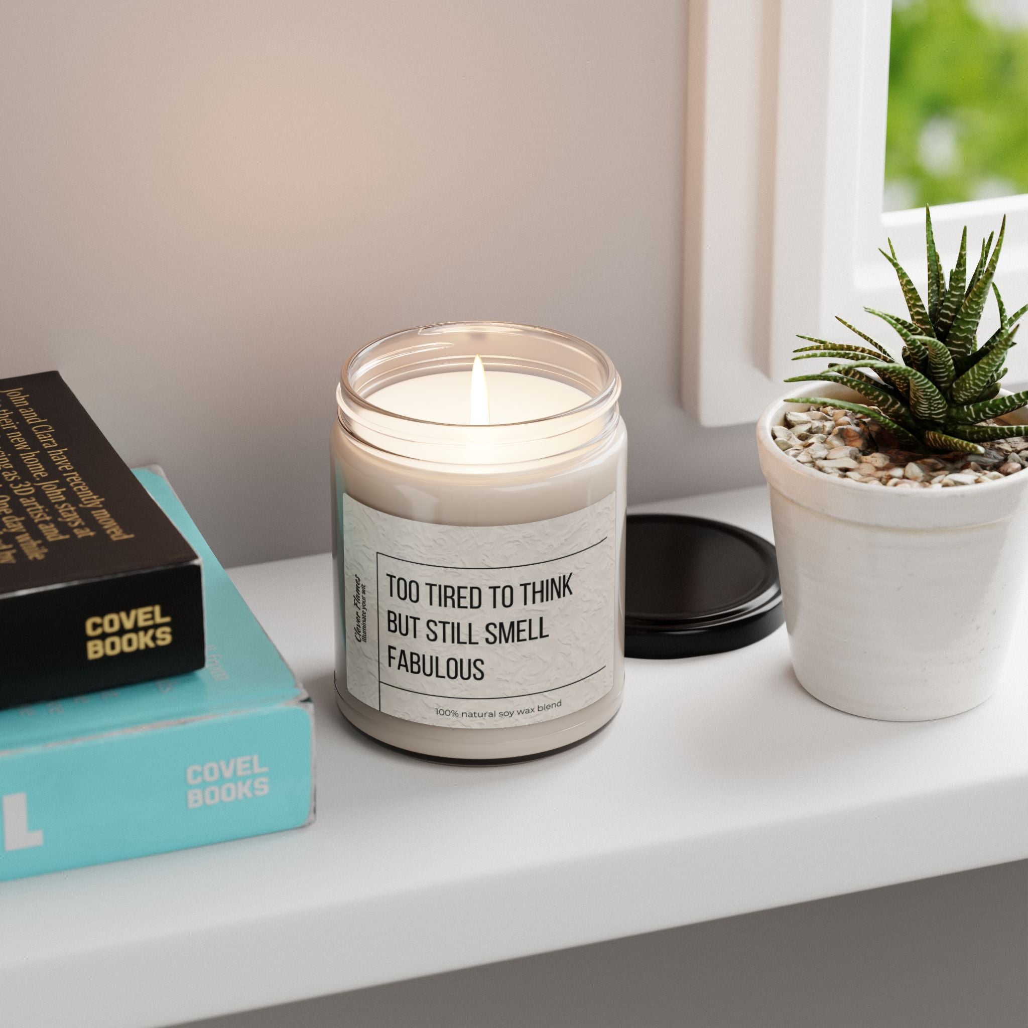 A soy candle from Printify, branded as Too Tired to Think Candle - Smells Like Fabulousness, rests on a shelf adorned with books and a small potted succulent. Bright sunlight streaming in through a nearby window bathes this eco-friendly setting, amplifying its calming aromatherapy allure.