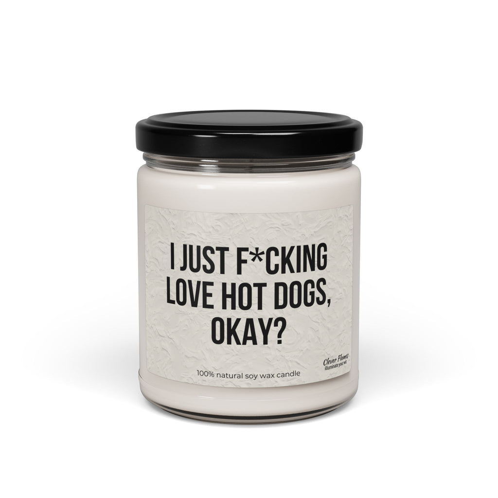The Printify "I Just F*cking Love Hot Dogs Okay?" Candle is a 9oz white, non-toxic scented soy candle in a reusable glass jar with black lid, featuring a label that reads the product name. Made from 100% natural soy wax.