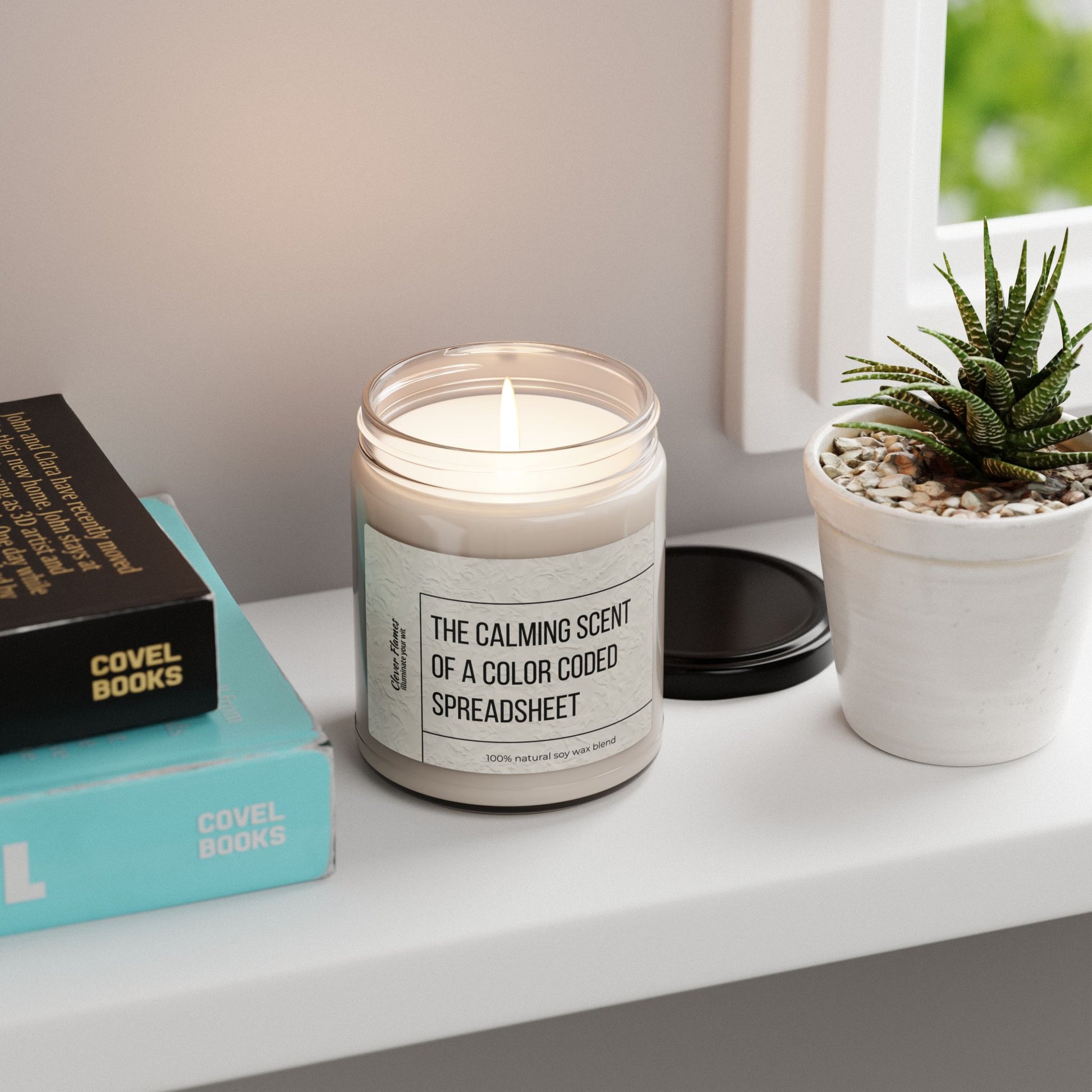 The Scented Soy Candle - The Calming Scent of a Color Coded Spreadsheet by Printify rests on a white shelf beside two books, a black book, and a small potted succulent—ideal for eco-friendly home fragrance lovers.