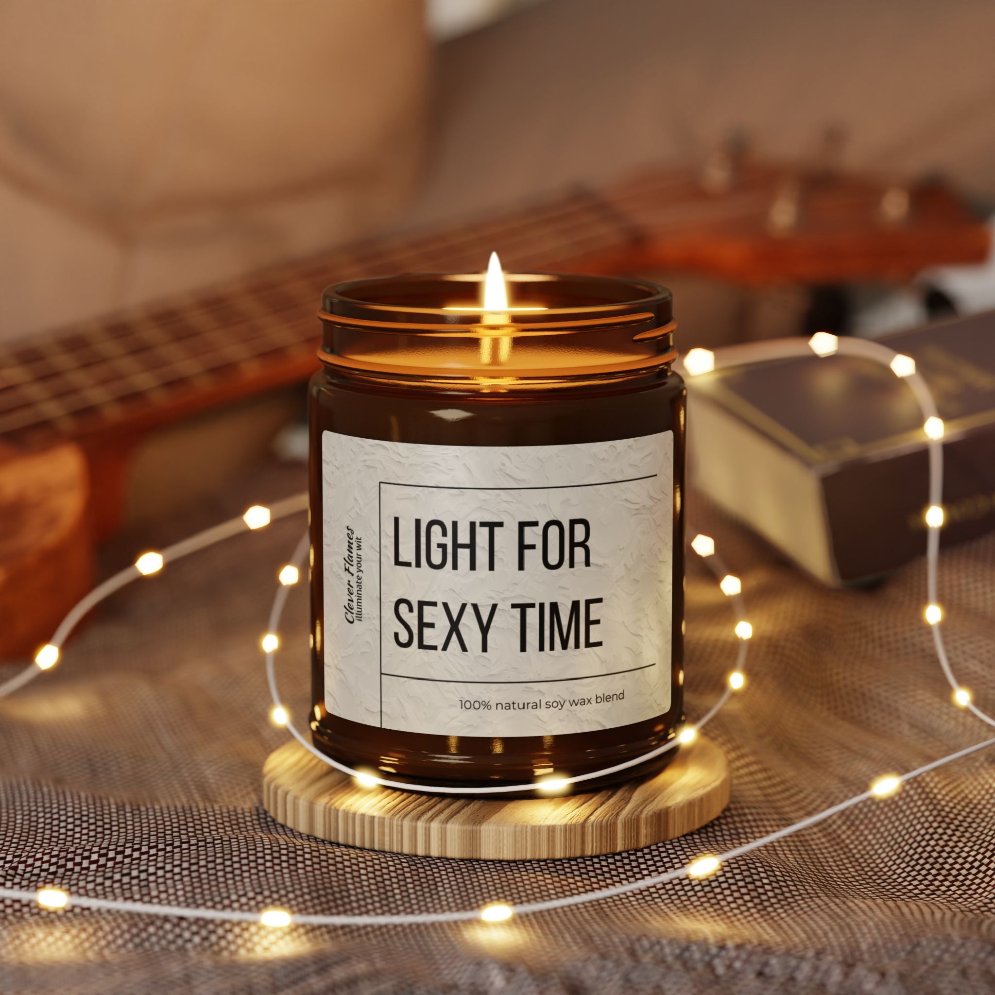 A lit brown Light for Sexy Time candle by Printify casts a warm glow on a wooden base. Surrounded by tiny lights with a blurred backdrop of a guitar and box, it creates an ideal cozy scene, perfect for anyone seeking unique raunchy gifts that add allure to any moment.