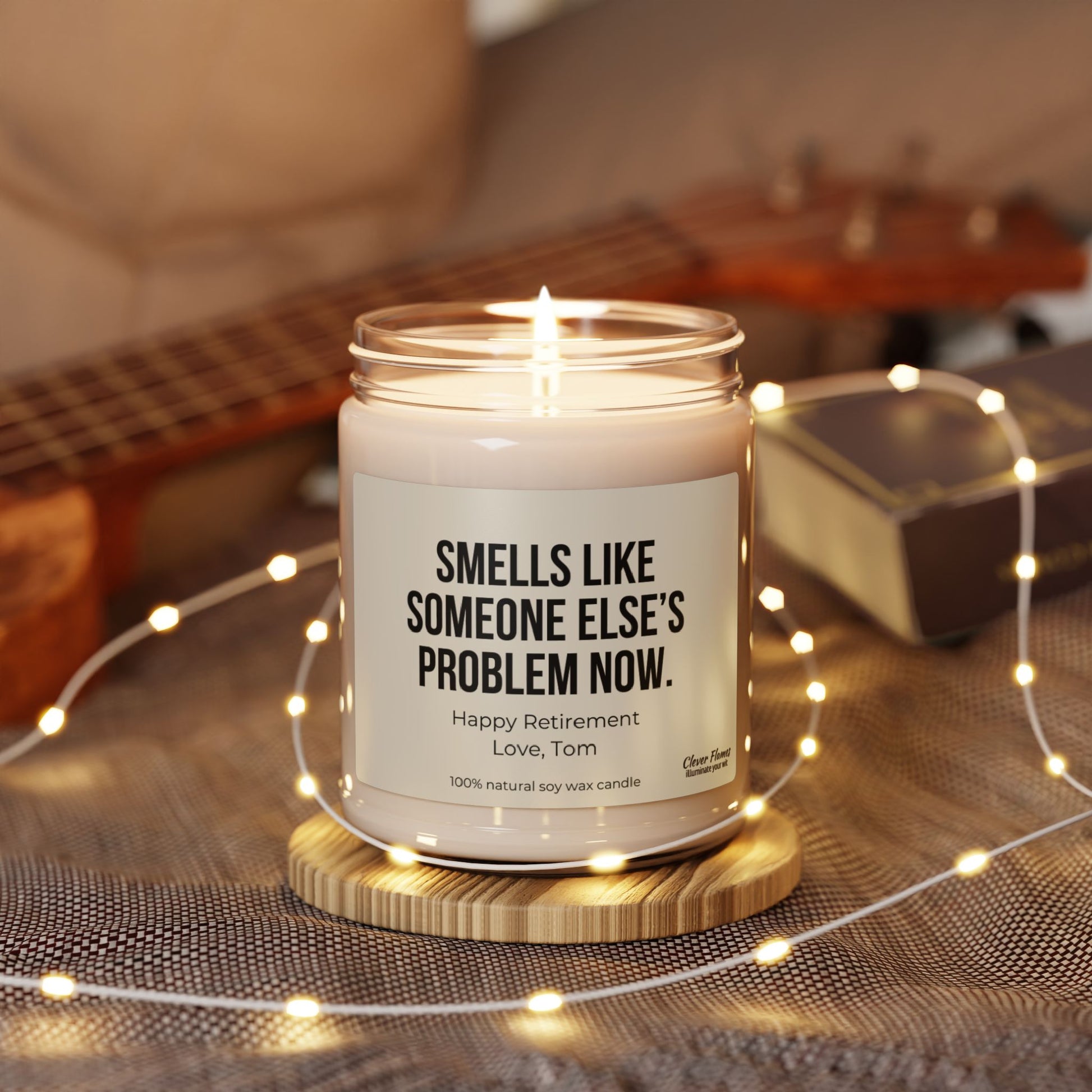 A personalized candle from Printify rests in a jar on a wooden coaster surrounded by string lights. The label reads, SMELLS LIKE SOMEONE ELSE’S PROBLEM NOW with the message "Happy Retirement Love, Tom." Made from 100% natural soy wax, this candle is the perfect humorous retirement gift. In the background, a ukulele is blurred.
