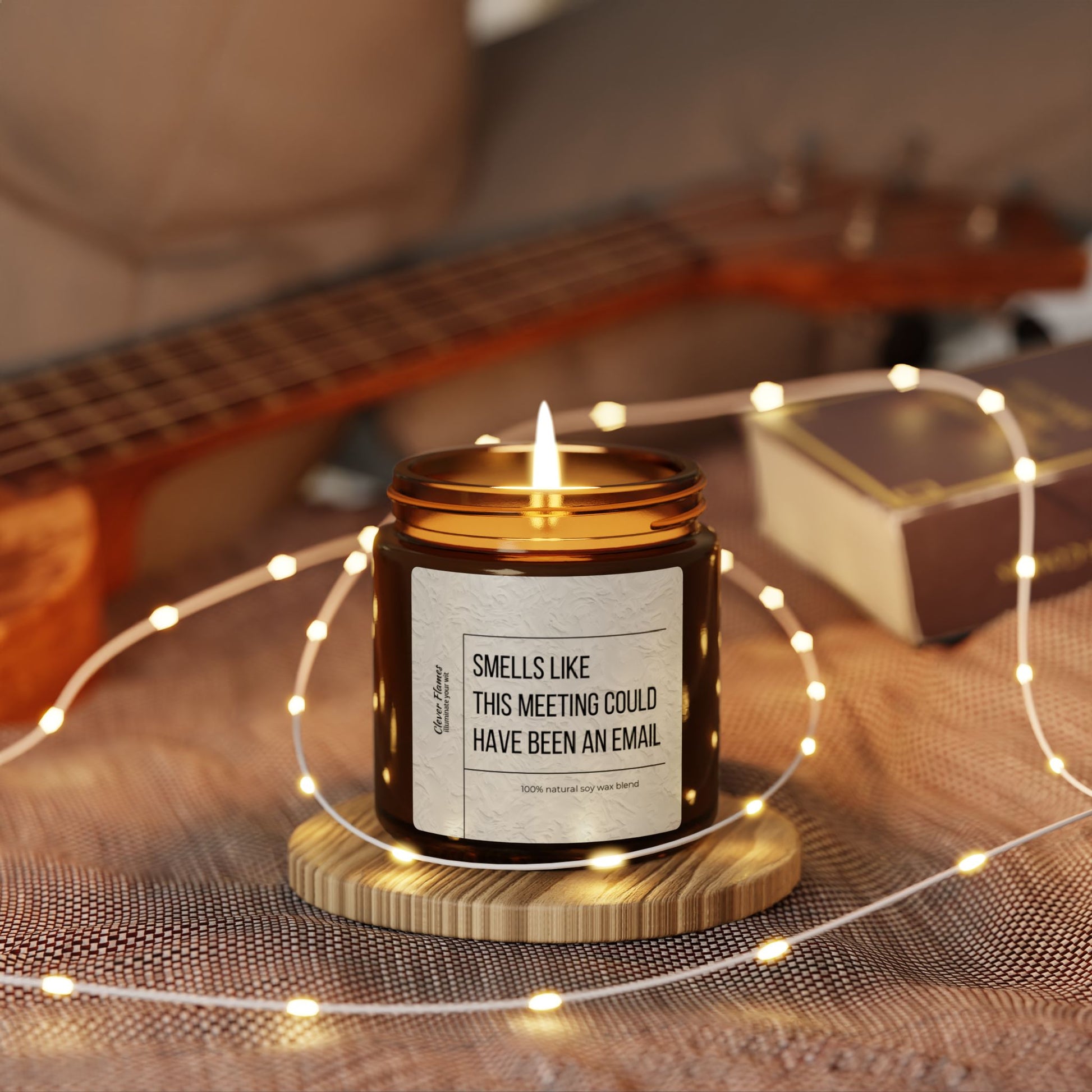 A Printify candle, branded as "Funny Office Candle - Smells Like This Meeting Could Have Been an Email," rests on a wooden coaster. Enveloped by string lights, with a guitar and box softly blurred in the background, this scented soy candle offers a cozy ambiance, making it the perfect gift for coworkers, bosses, or those who work from home.