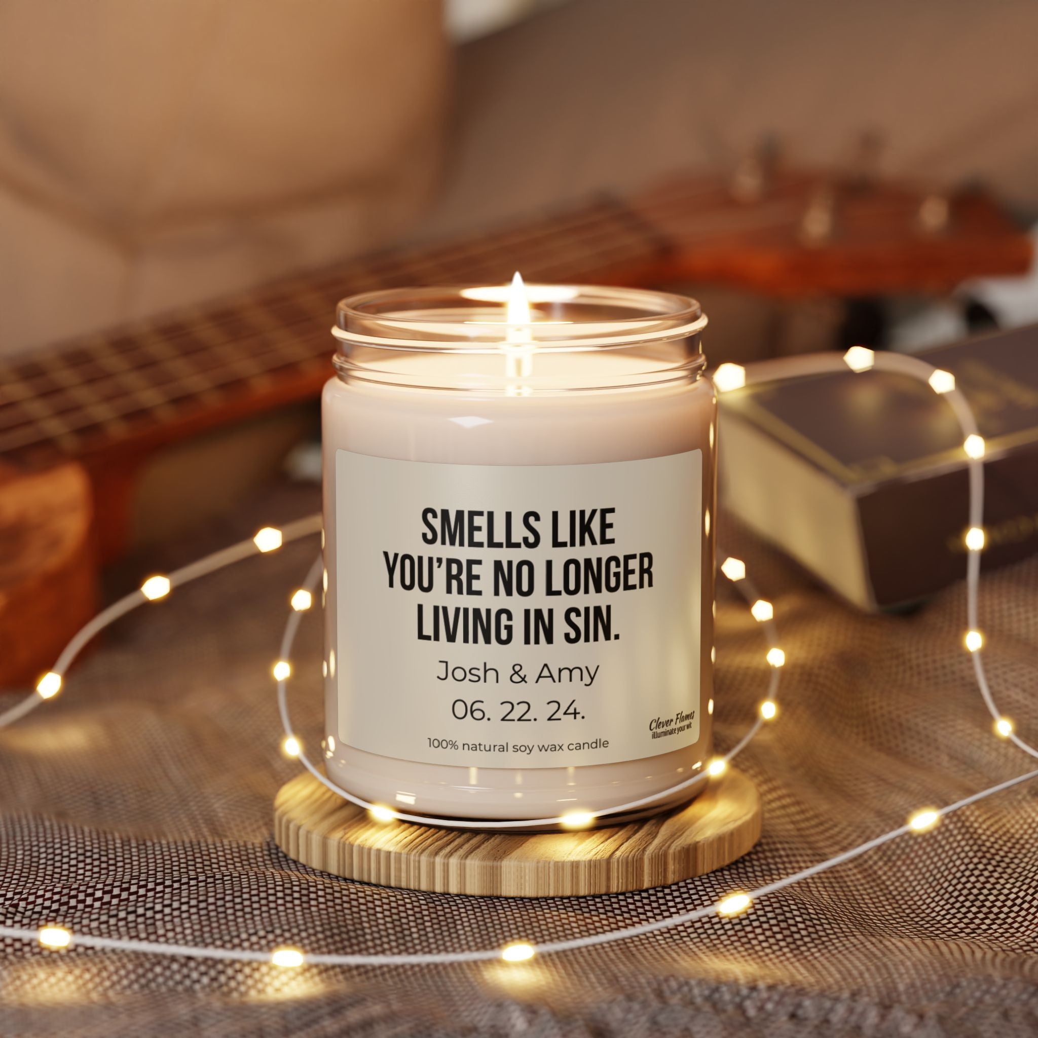A lit No Longer Living In Sin Soy Candle from Wedding is displayed in a glass jar on a wooden coaster, encircled by string lights. The label says, "Smells like you're no longer living in sin. Josh & Amy 06.22.24." This 100% natural scented candle, featuring a guitar resting softly in the background, makes a perfect engagement gift for couples.