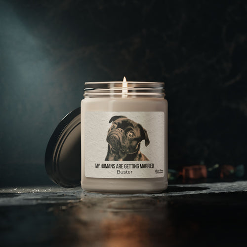 The My Humans Are Getting Married Personalized Pet Engagement Candle by Printify features a black dog with custom text, making it a thoughtful engagement gift. The lit white candle comes in a glass jar, with the lid beside it on a dark surface.