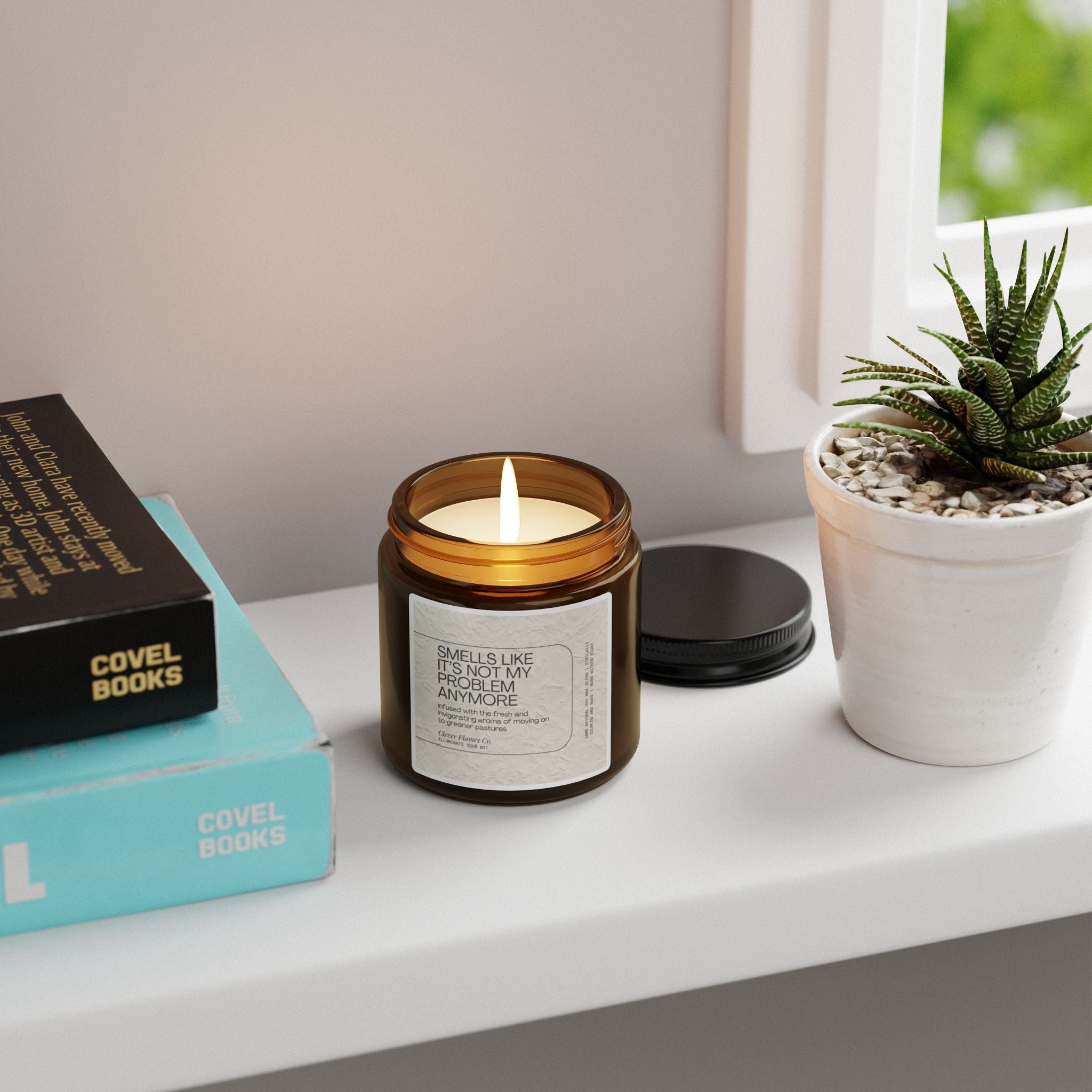 A lit soy candle, labeled with "Not My Problem Anymore," rests in a brown jar on the windowsill next to a small potted succulent and neatly stacked books. This scented candle, from Printify, features the Atlantis Whisper fragrance. The white window frame allows natural light to gently filter through.