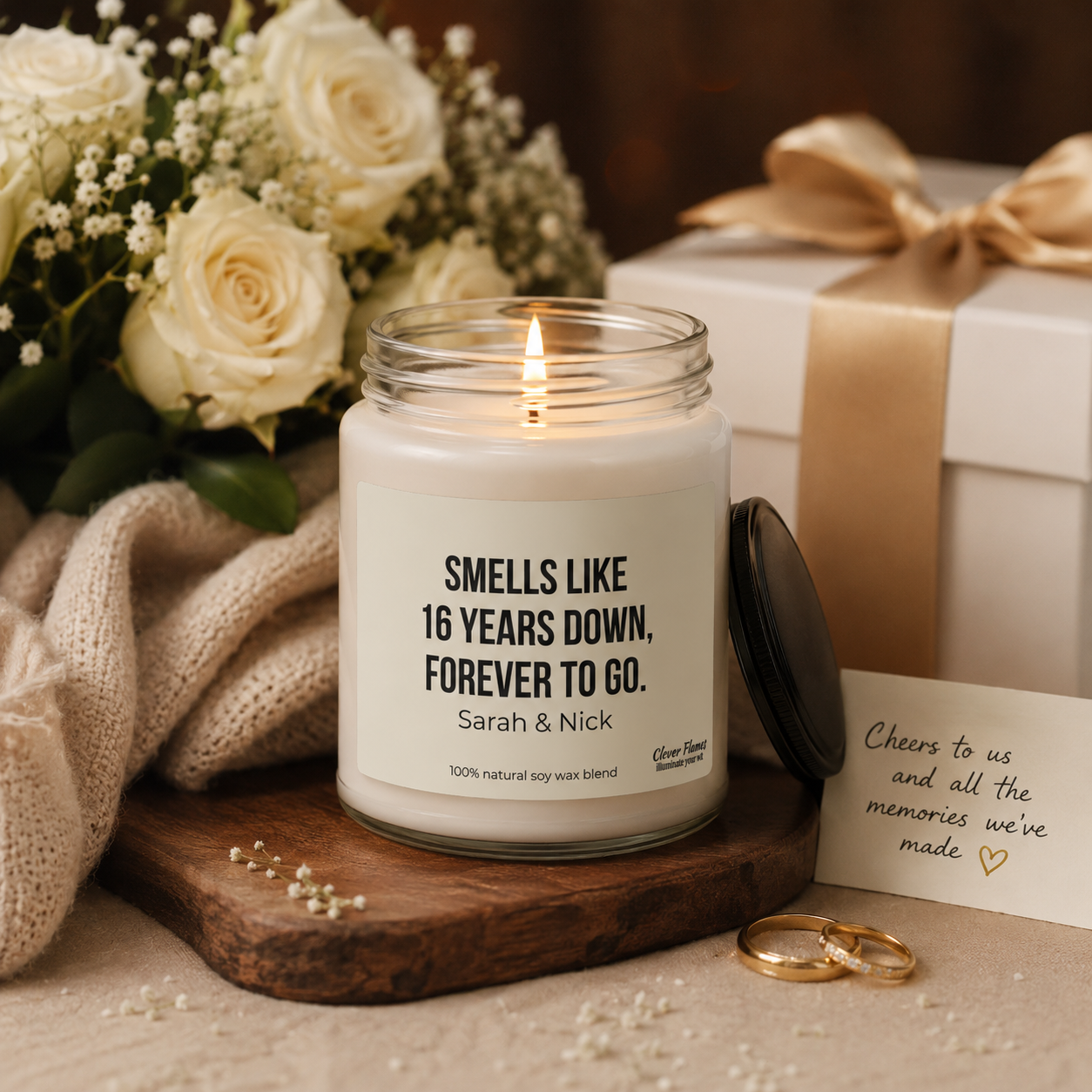 The Printify Personalized 16 Years Down, Forever to Go Candle – Wax Anniversary Gift for Couples sits on a wooden tray, paired with gold rings, white roses, a note, a knitted scarf, and a wrapped gift.